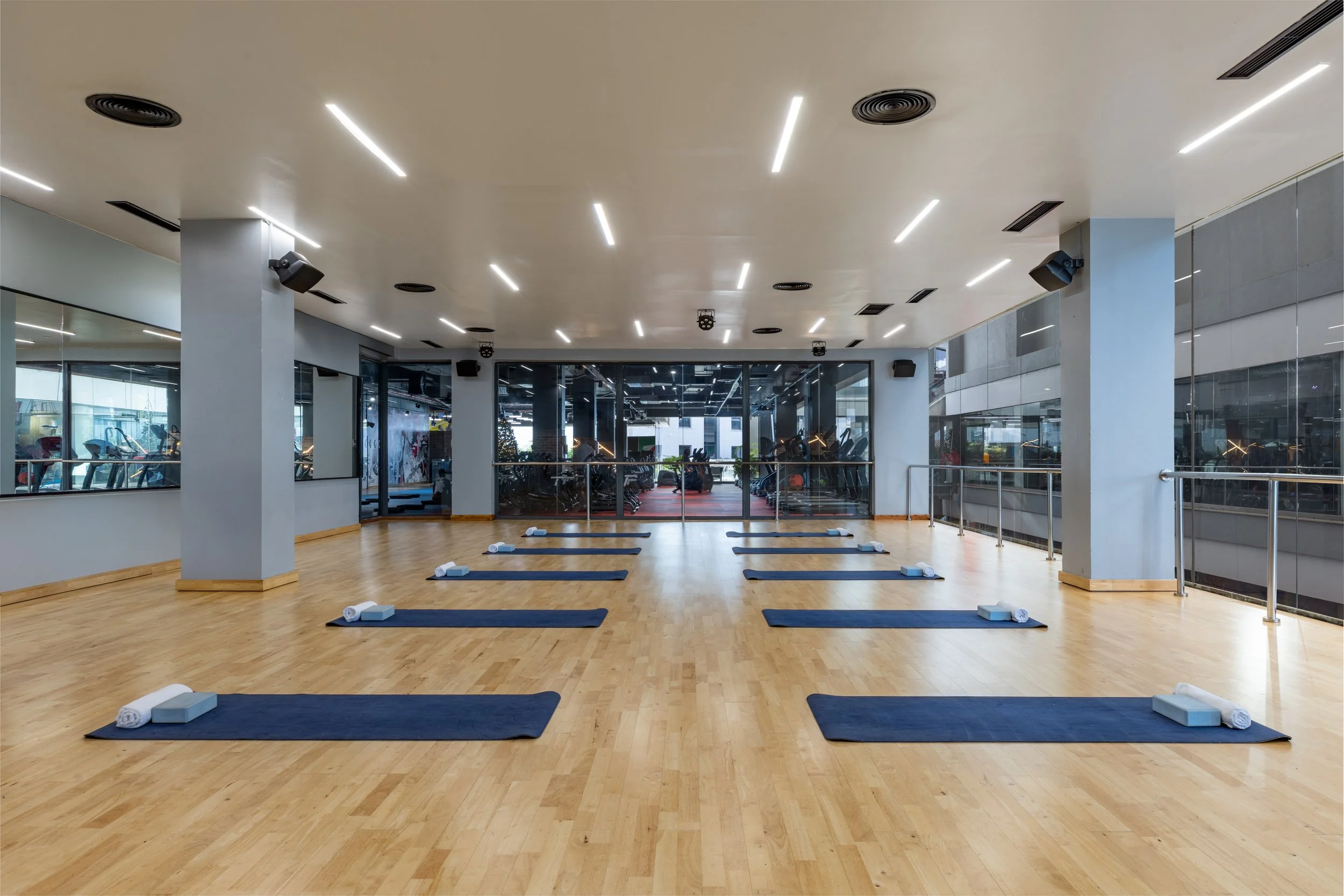 Empty yoga or fitness studio with blue mats and foam blocks arranged in a pattern, large windows, mirrored wall, and wooden floors.
