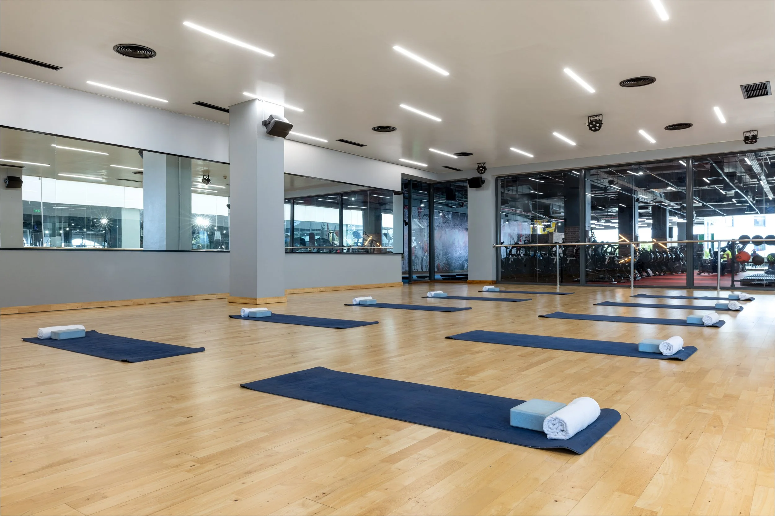 Empty yoga studio with black mats and rolled towels on a wooden floor, mirrored wall, and large glass windows showing a gym area outside.