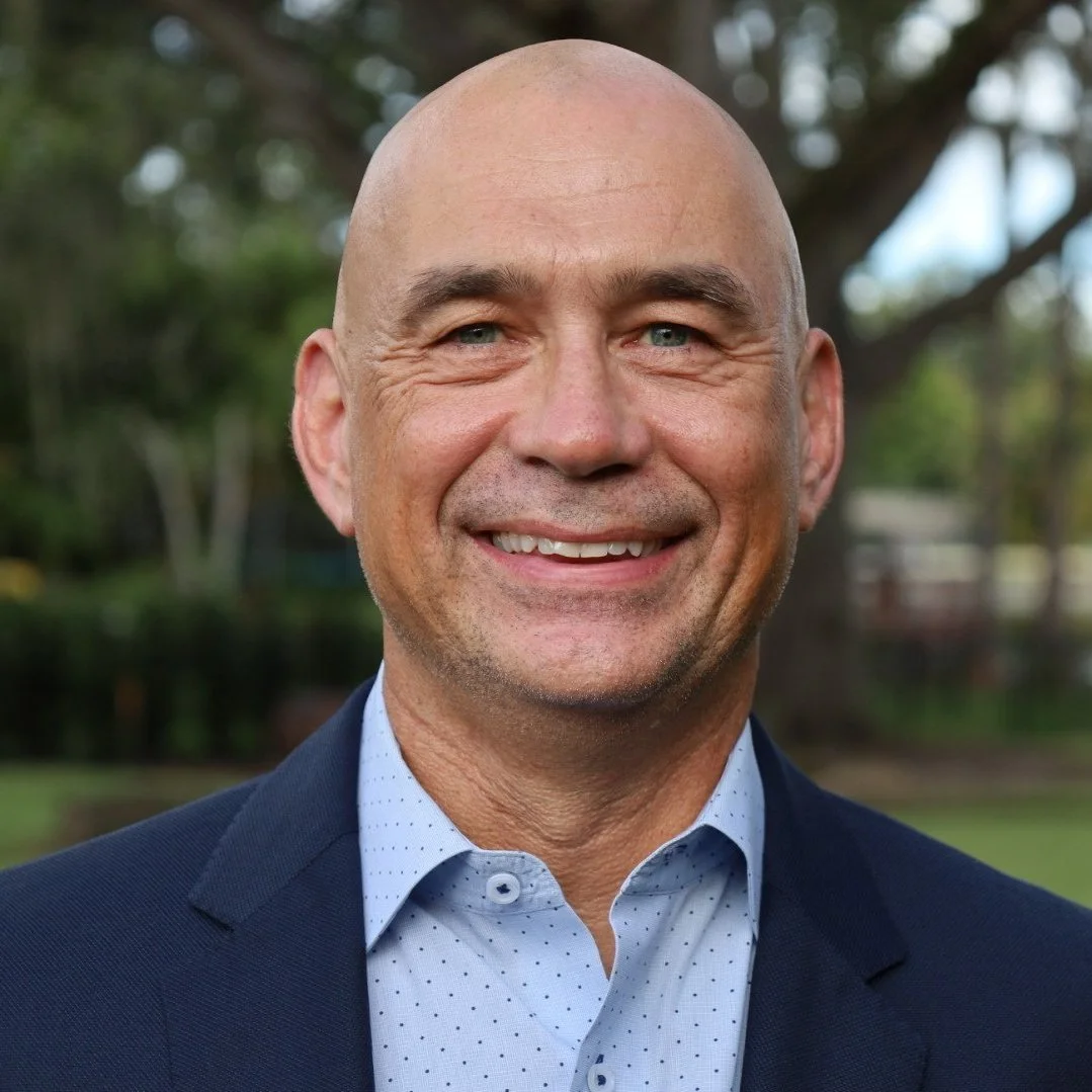 Smiling middle-aged man with a shaved head wearing a navy blazer and light blue shirt outdoors with trees in the background.