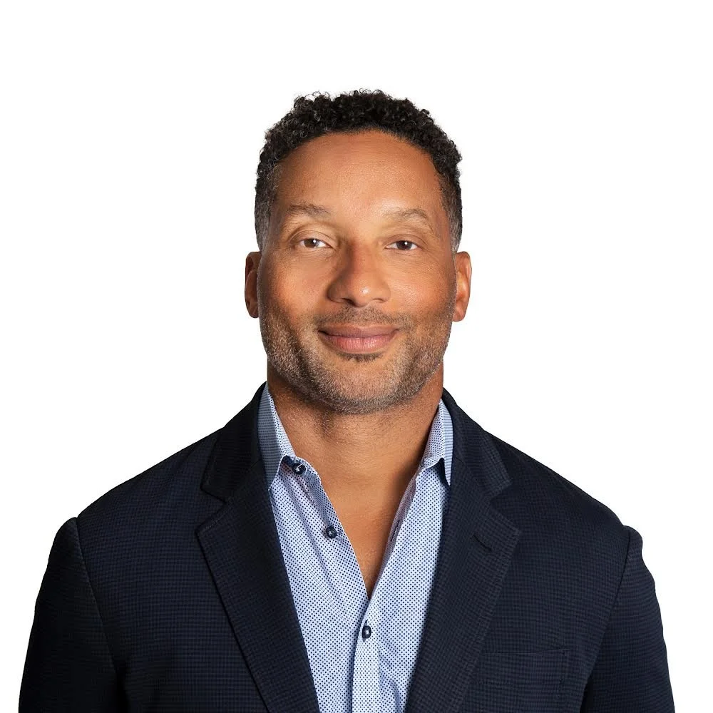 Headshot of a smiling man with short, curly dark hair, wearing a dark blazer and patterned shirt, against a white background.