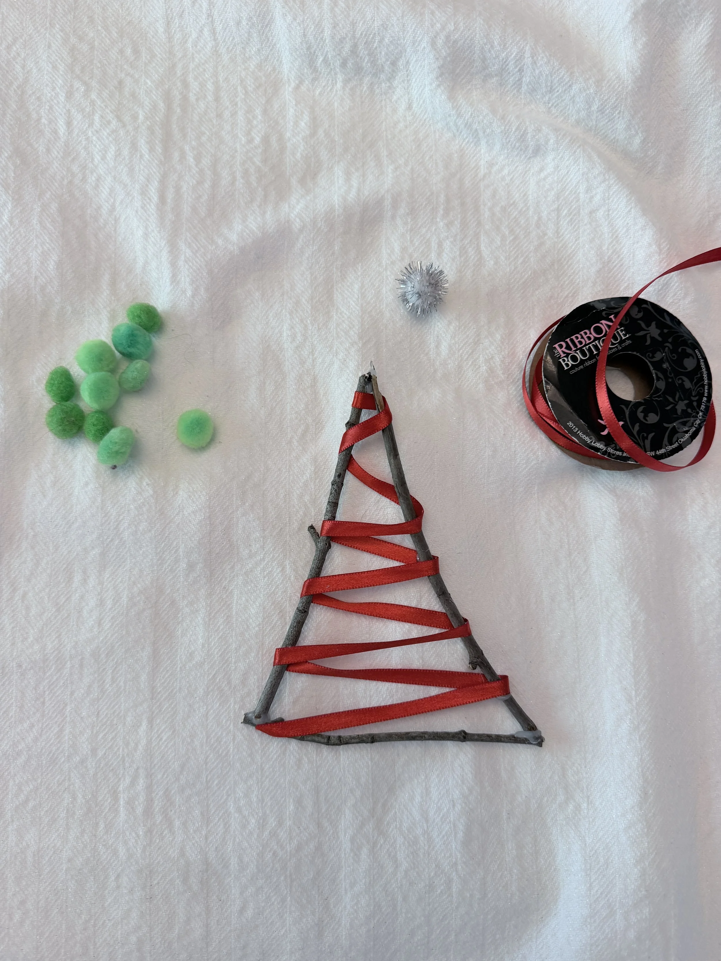 Christmas tree shaped craft made of three twigs glued together with red ribbon wrapped around it and a pile of green pom pom decorations off to the side.
