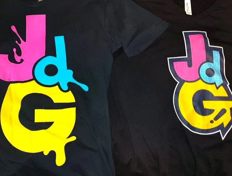 Two black T-shirts with colorful graffiti-style logos featuring the letters 'JdG' in pink, blue, and yellow.