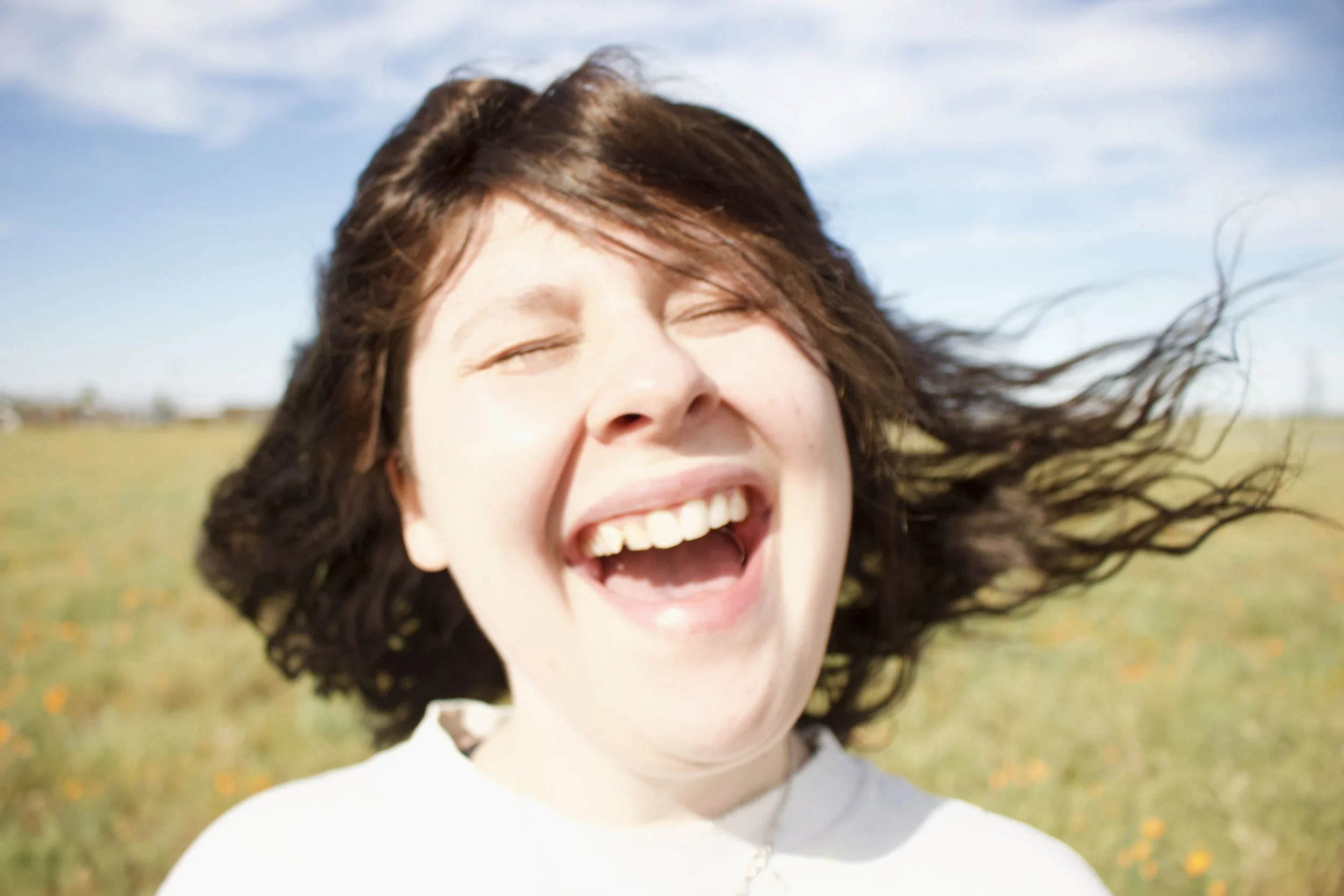 A woman with brown hair smiling and laughing outdoors in a field during daytime, with her hair blowing in the wind.