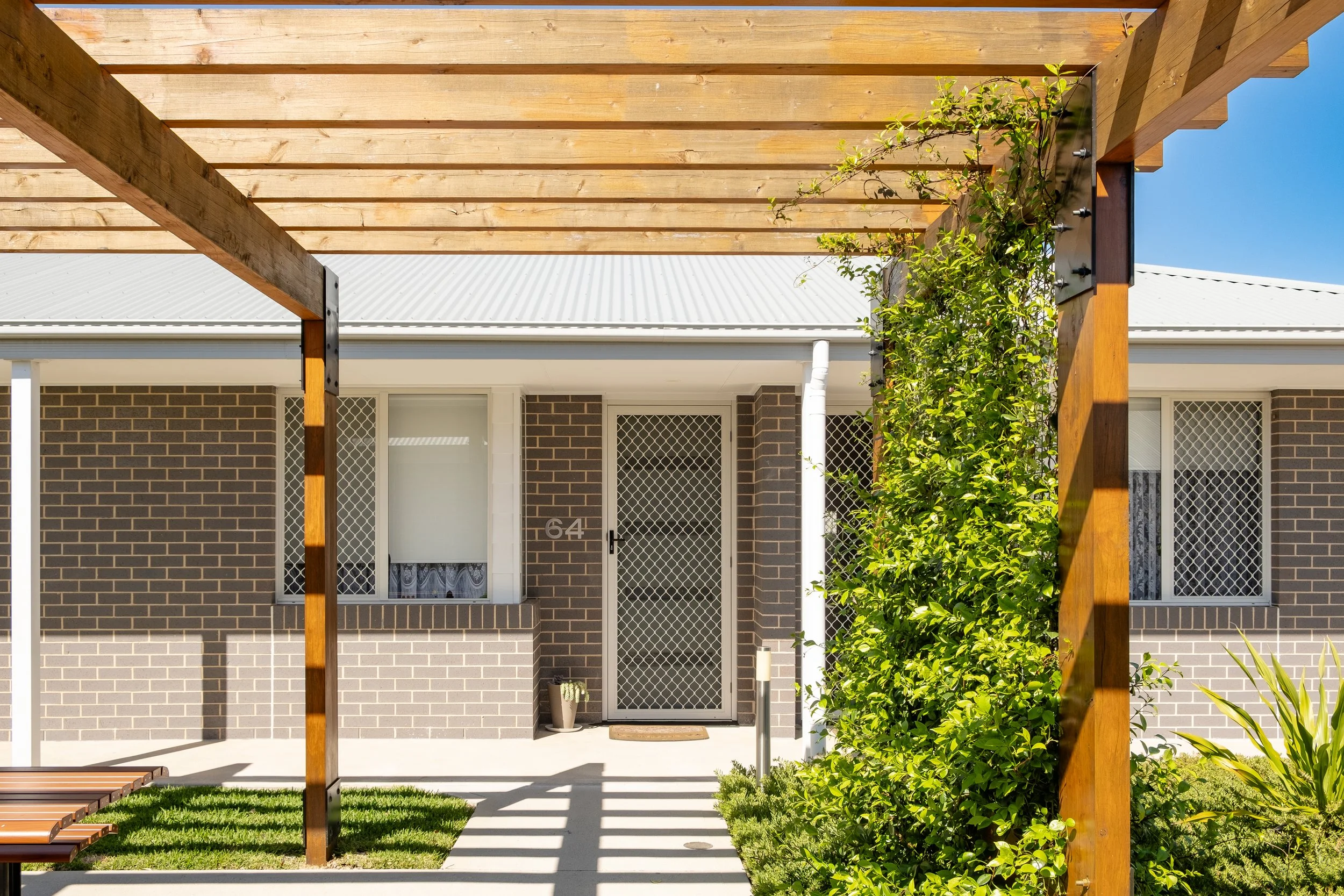View of a row of over 55's villas with screened doors, brick walls, and a covered walkway, surrounded by green grass and a small tree.