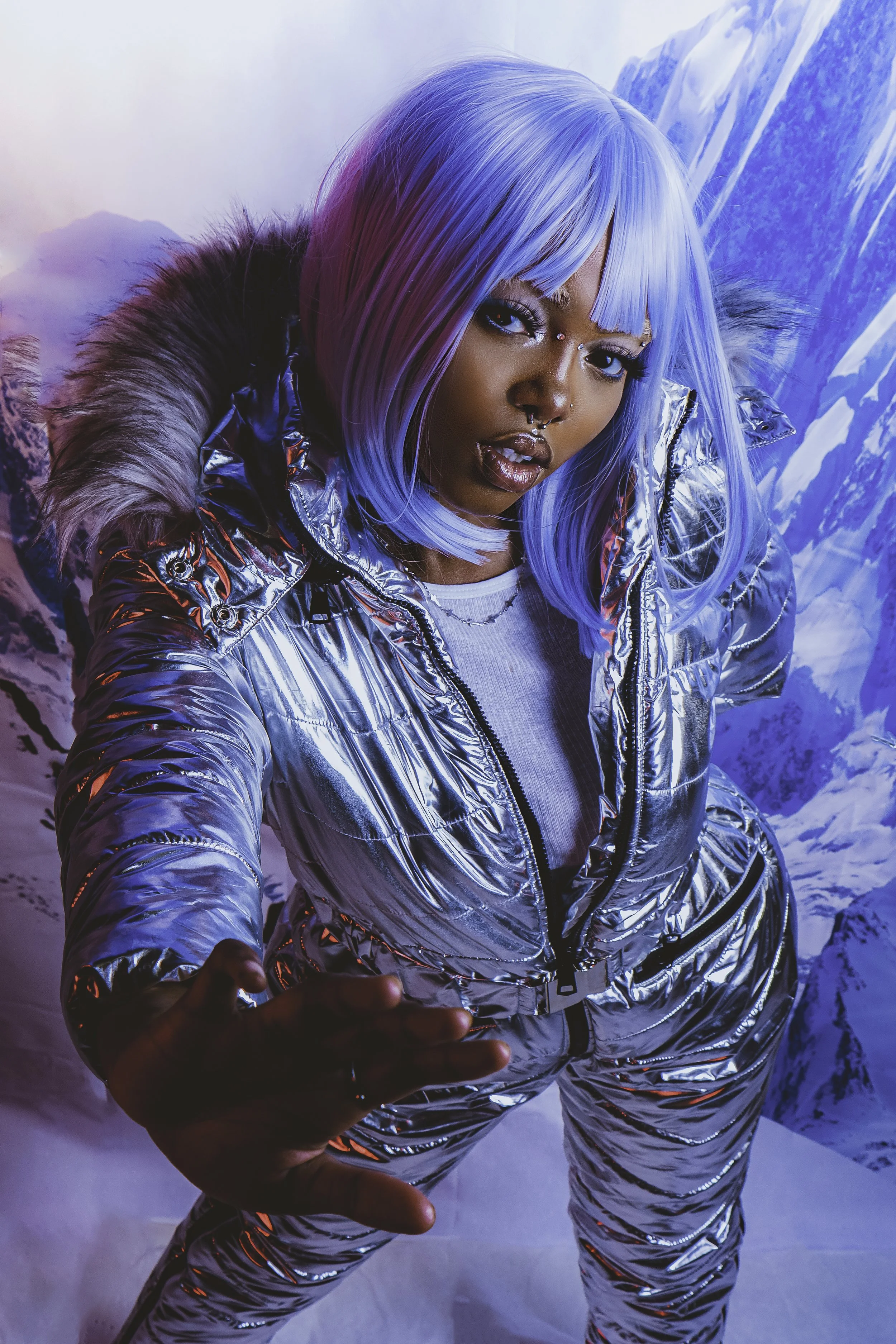 A woman with light blue hair, wearing a shiny silver puffer jacket, taking a selfie in a snowy mountain landscape.