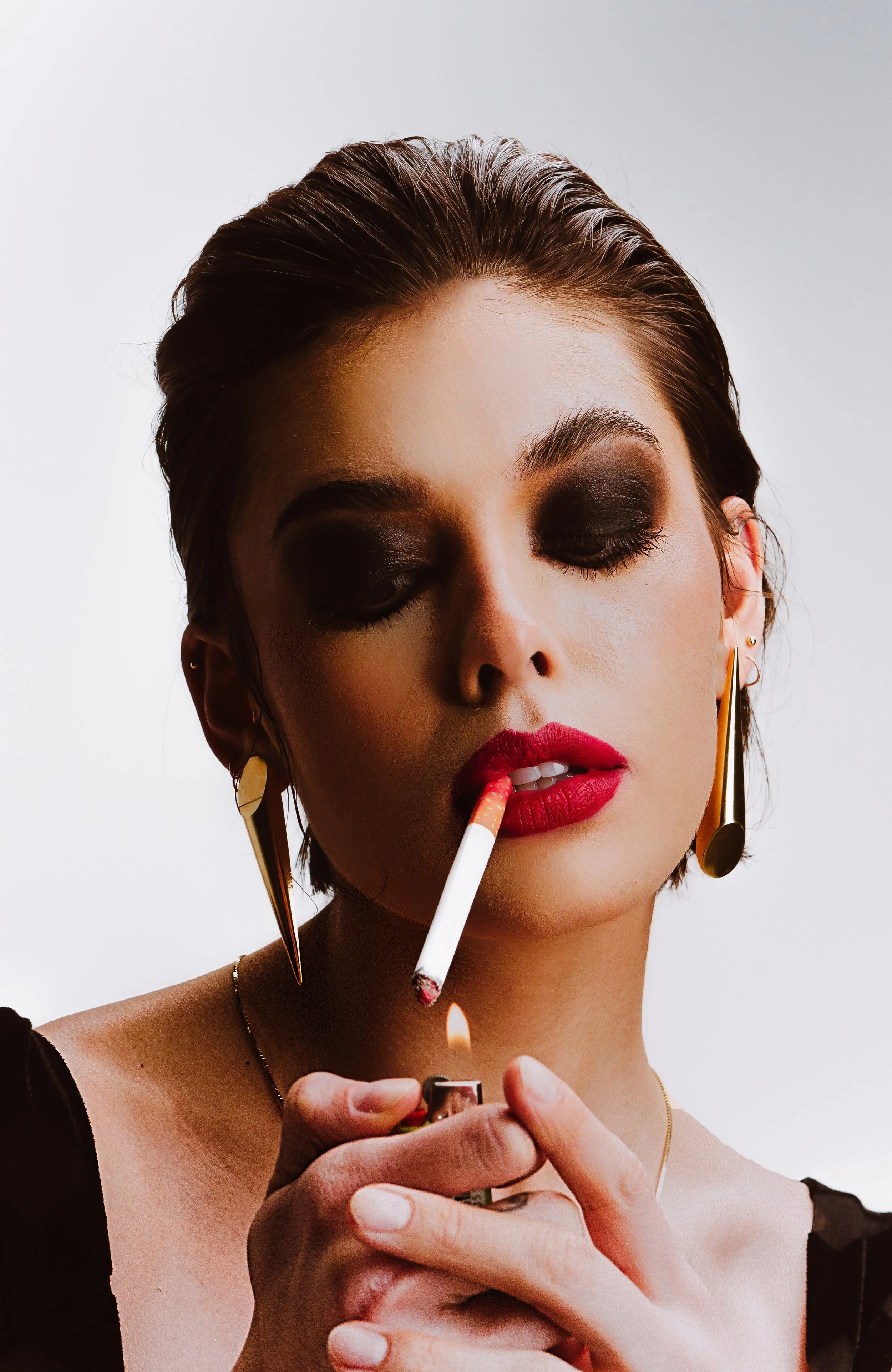 A woman with dark, styled hair, wearing bold makeup and gold earrings, is lighting a cigarette with a match.