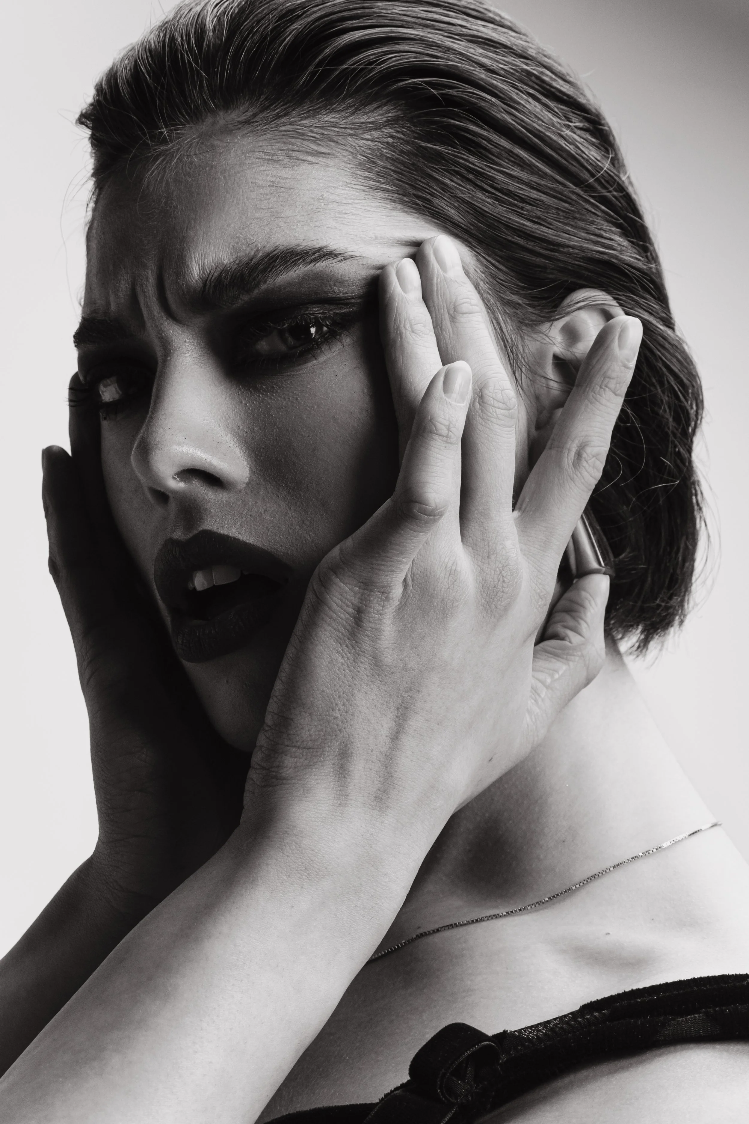 A black and white photo of a woman with one eye closed, her mouth slightly open, and her hand covering part of her face. She has wavy hair and is wearing a thin chain necklace.