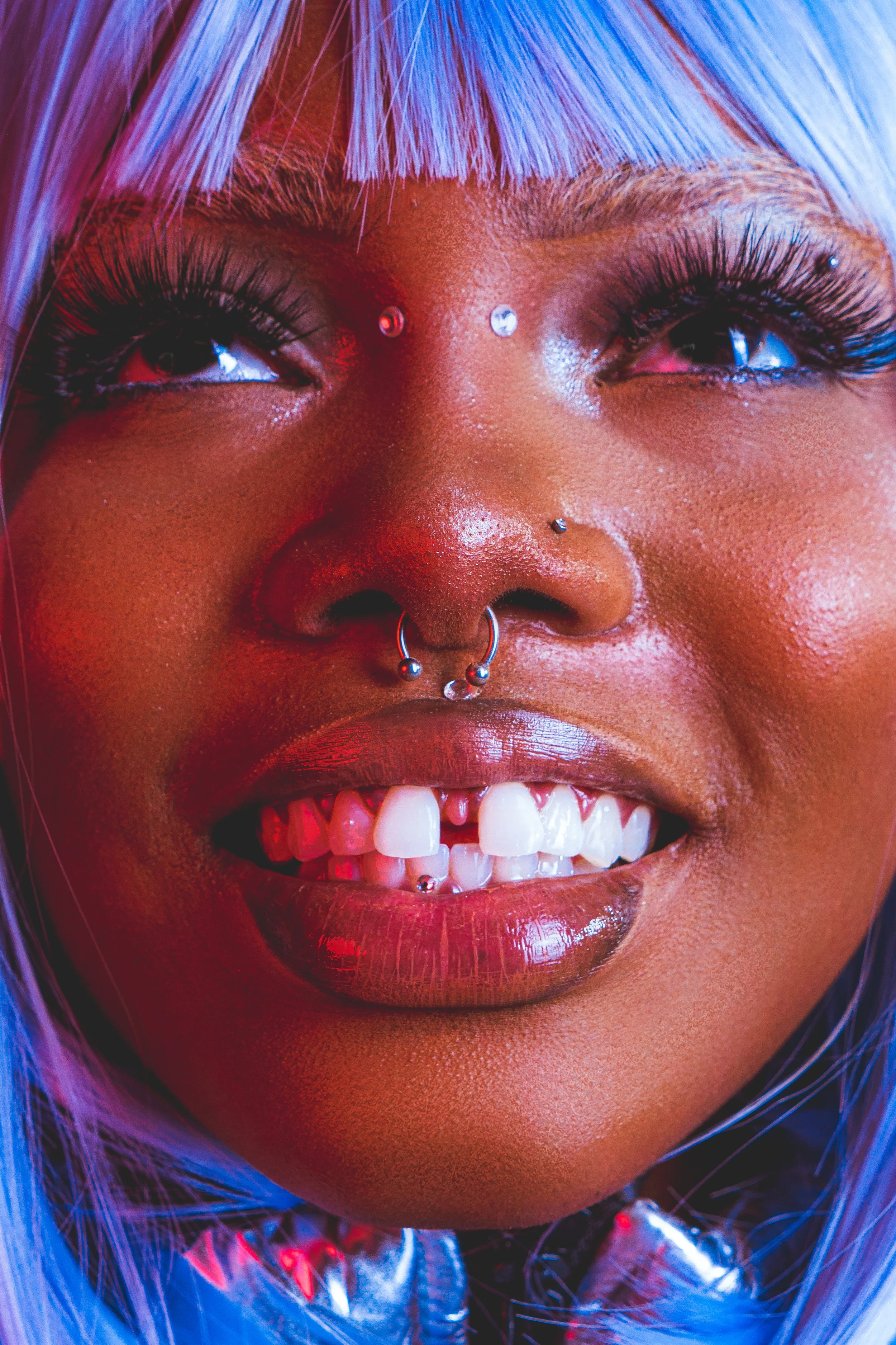 Close-up of a smiling woman with blue and purple hair, multiple facial piercings including a nose ring, septum piercing, and eyebrow studs, and wearing dramatic makeup with long eyelashes.