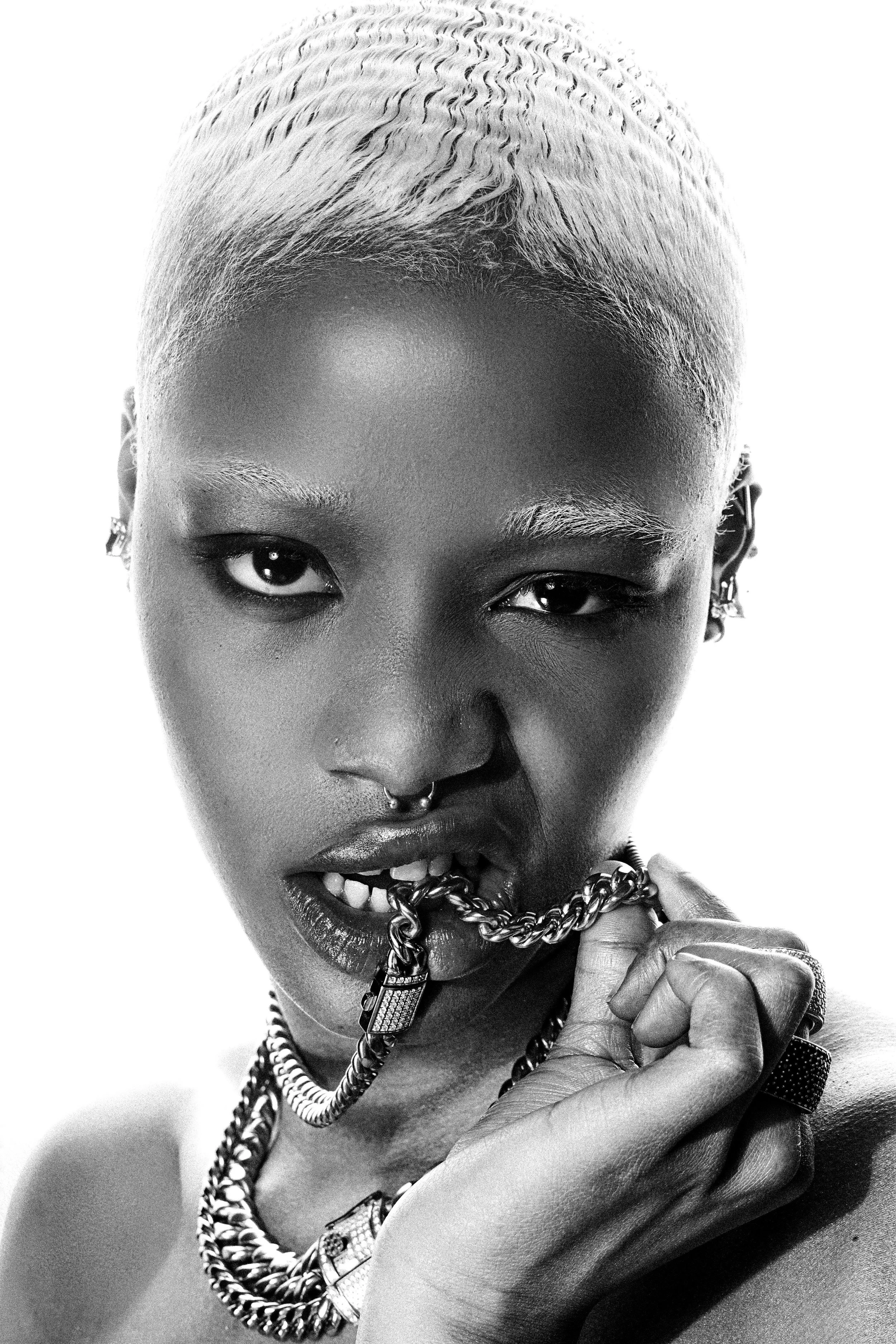 Close-up black and white portrait of a woman with platinum blonde hair, multiple earrings, wearing layered chains, holding a chain necklace close to her lips, with a confident expression.