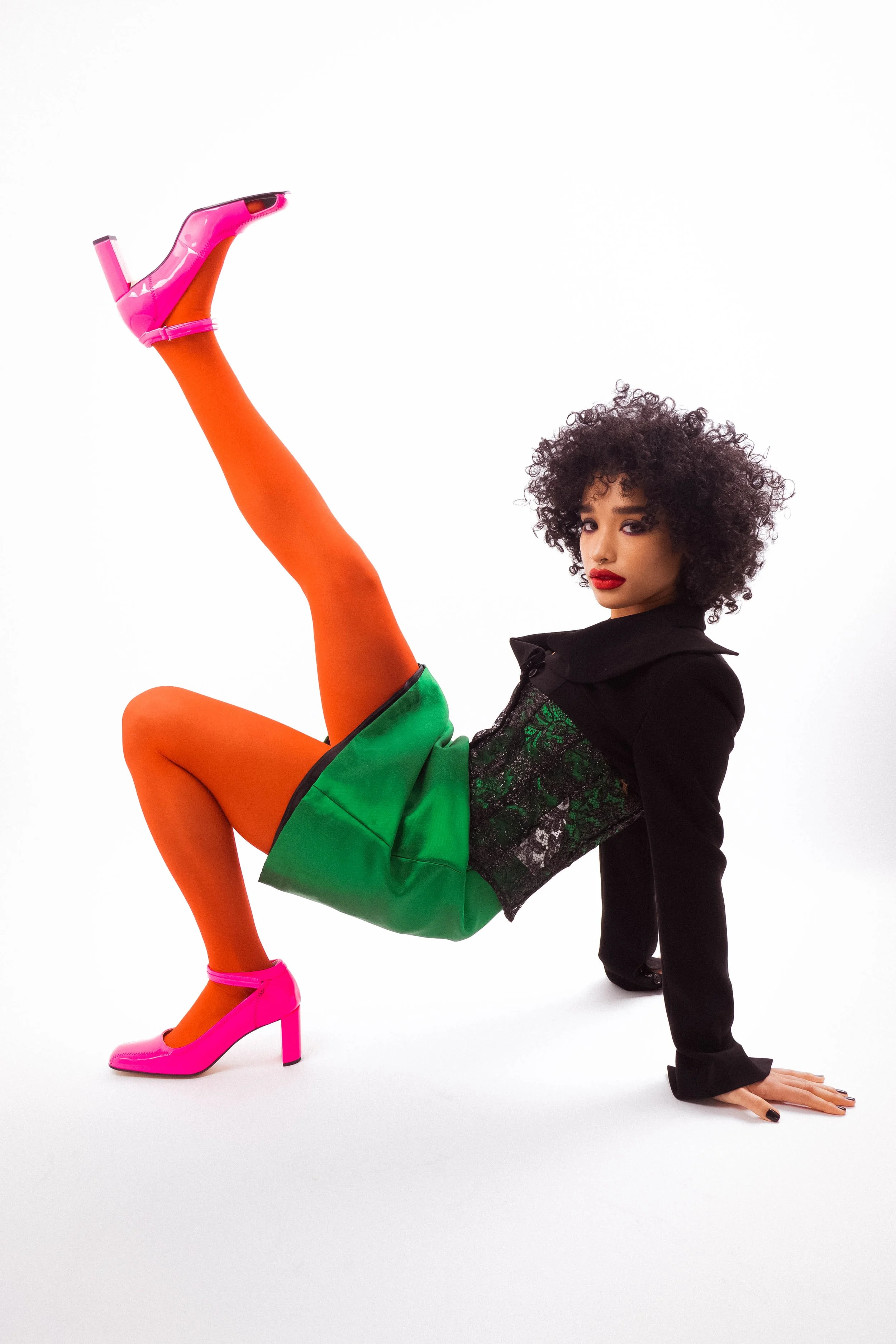 A woman in a black jacket with green lace details, orange tights, and bright pink high-heeled shoes is in a creative pose on a white background. She is supporting herself on her hands and feet, with one leg raised high in the air.