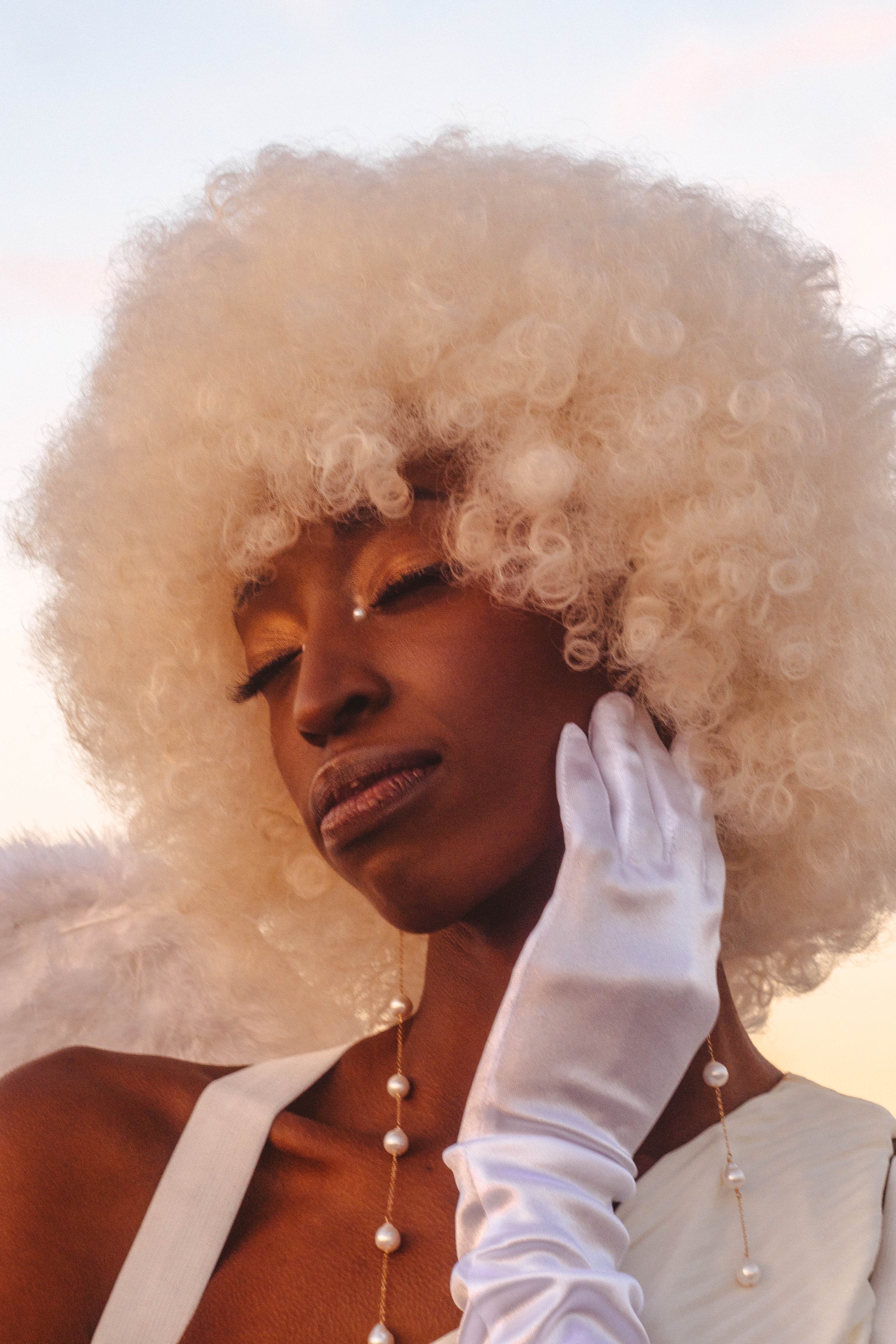 A woman with dark skin and platinum blonde, curly hair, wearing gold eyeshadow, a nose piercing, and pearl jewelry, dressed in white with a glove, posing against a light background.