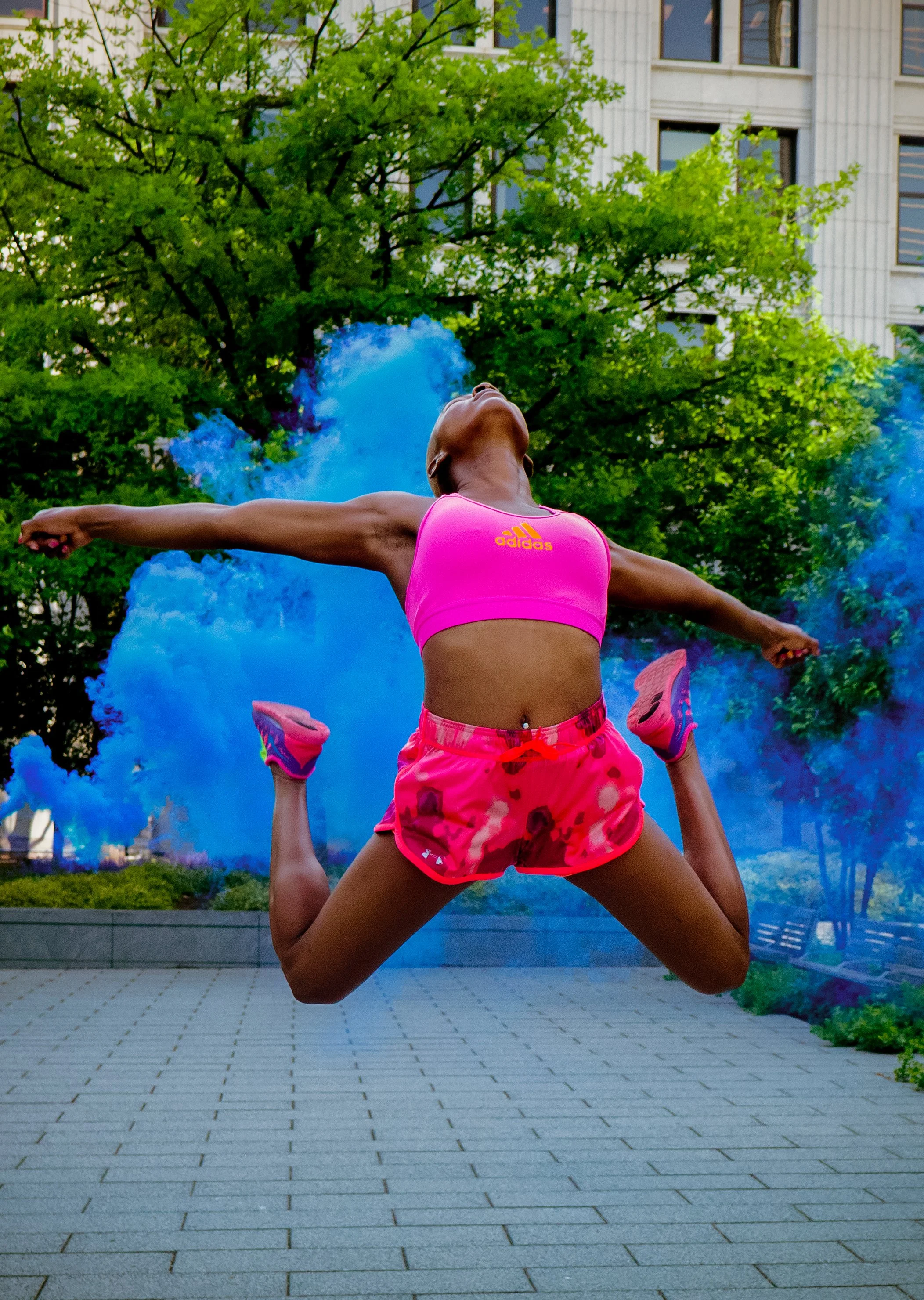 A woman in a pink sports top and shorts performing a jump in an outdoor urban setting with a person holding her legs, blue smoke, a tree, and a building in the background.