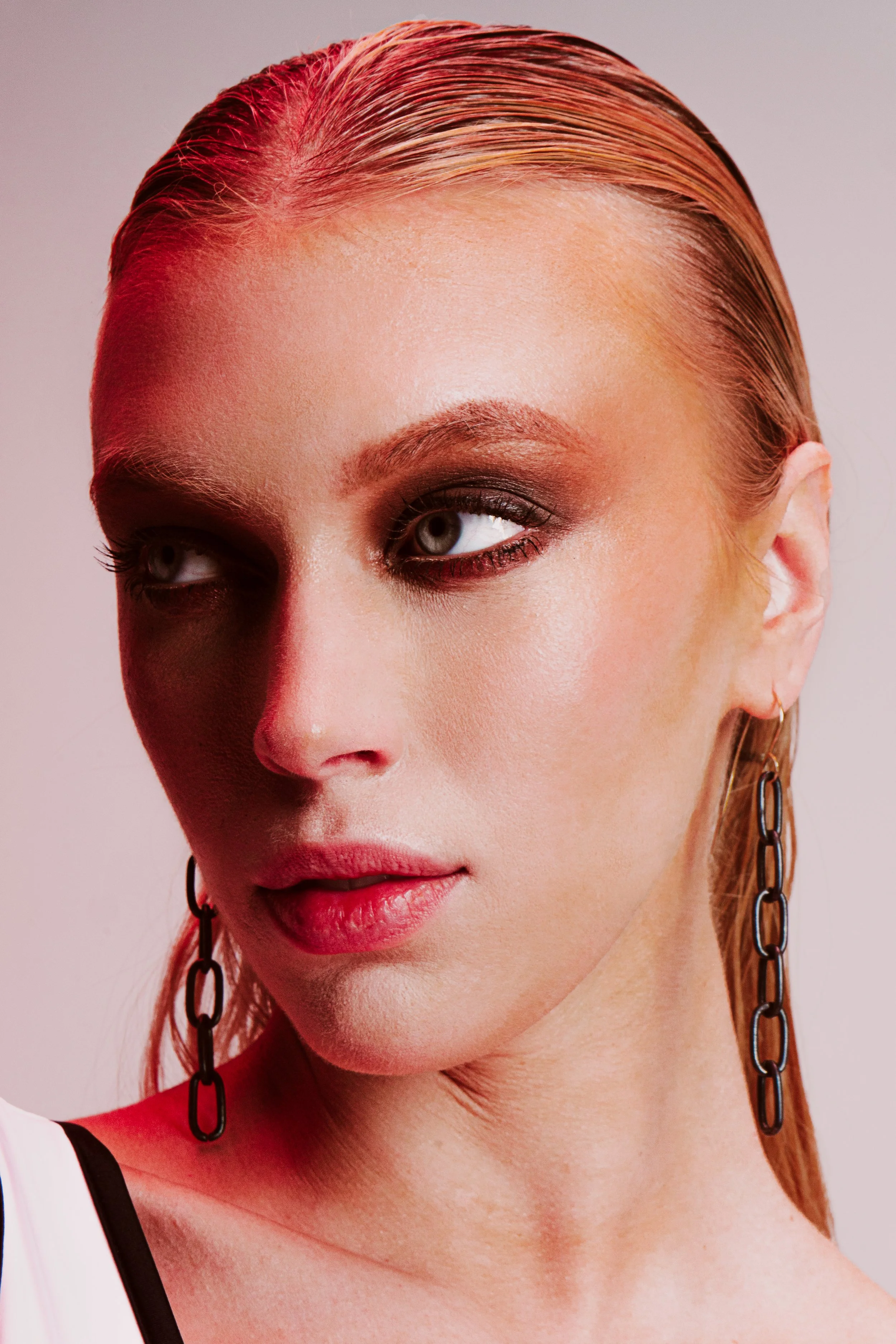 Close-up of a woman's face showing her left eye, part of her lips, and ear with a chain earring, with blond hair slicked back.