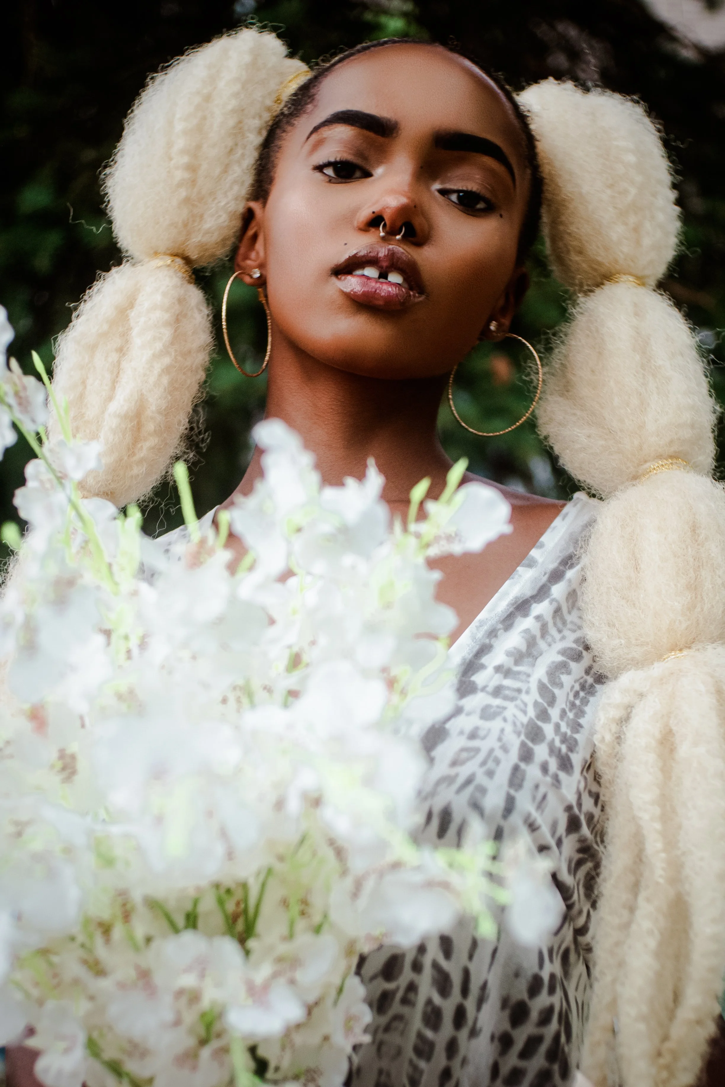 Close-up of a woman with tan skin, dark eyebrows, bold lipstick, wearing large hoop earrings and a septum piercing, posing outdoors with white flowers and greenery in the background.
