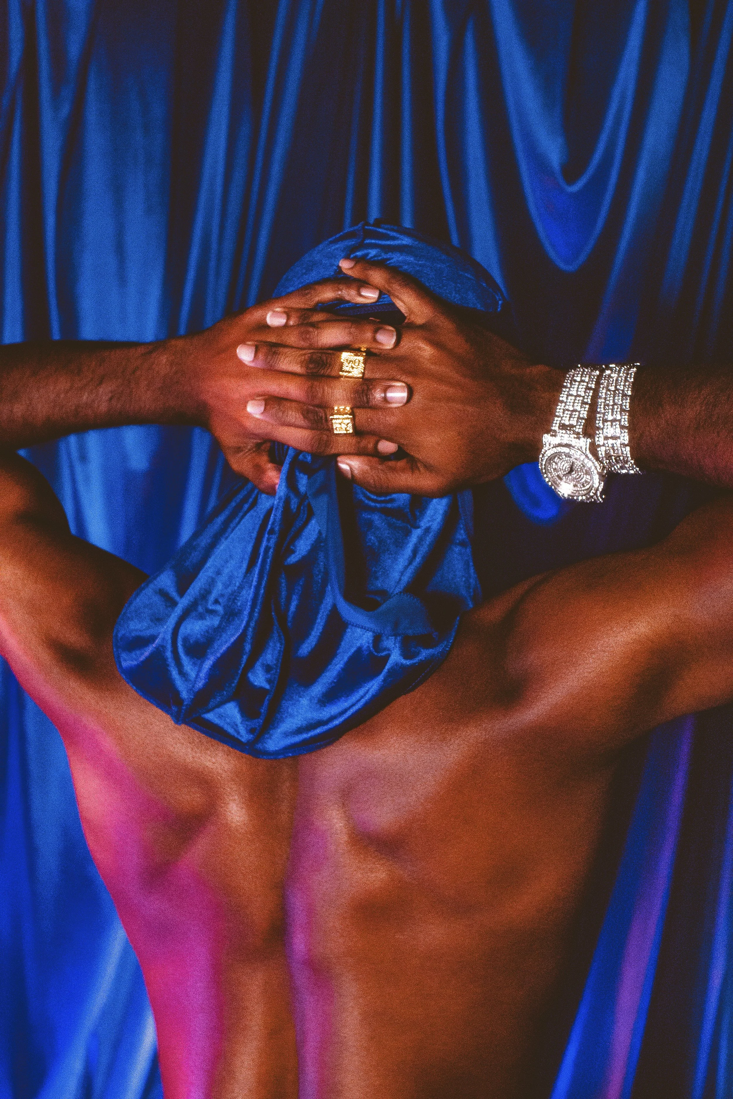 Close-up of a person's hands, adorned with gold rings and a silver watch, clasped over their thighs, with a deep blue fabric and draped backdrop in the background.