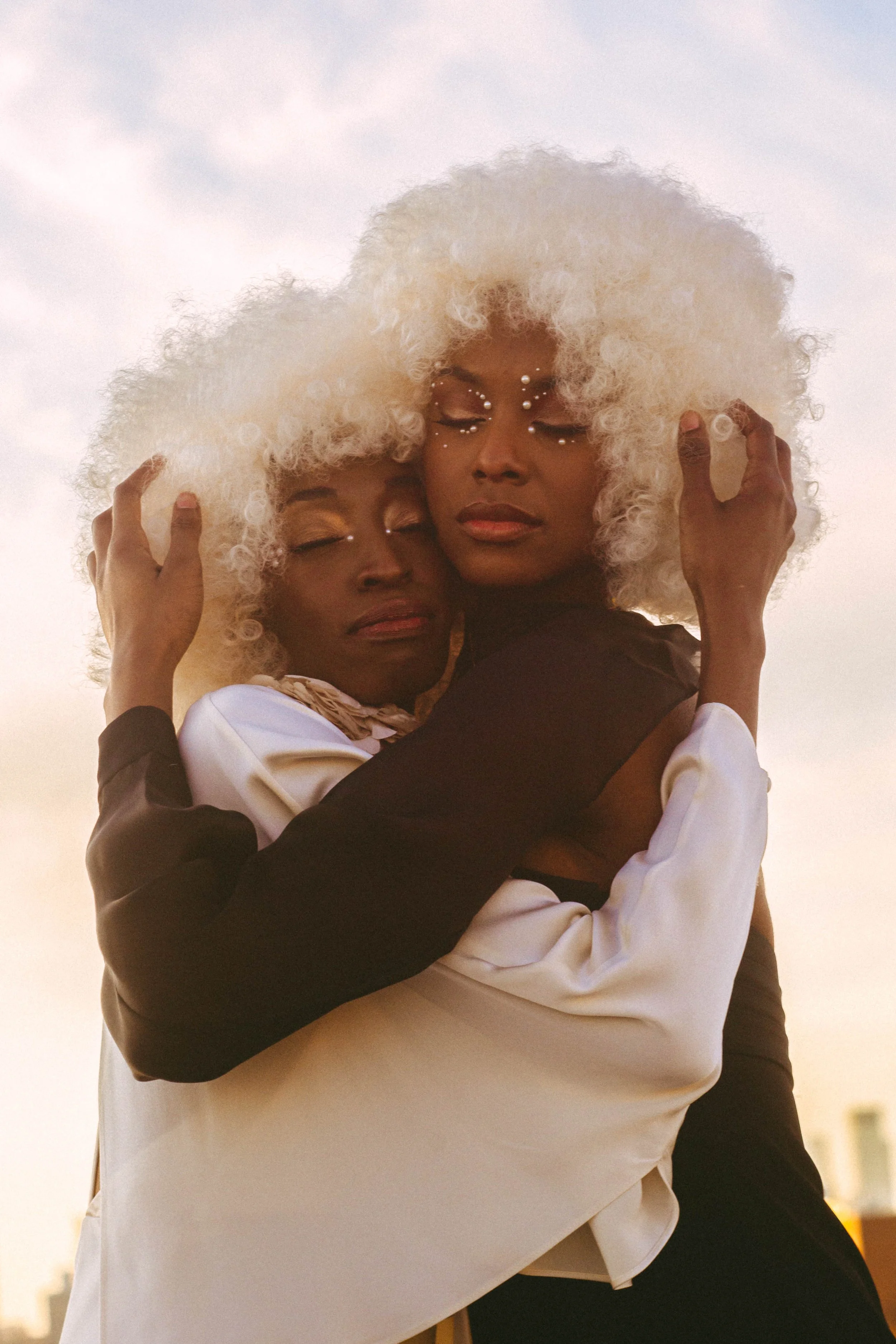 Two women with creative white curly hairstyles embracing each other, wearing makeup with rhinestones around their eyes, and dressed in contrasting black and white outfits against a soft sky background.