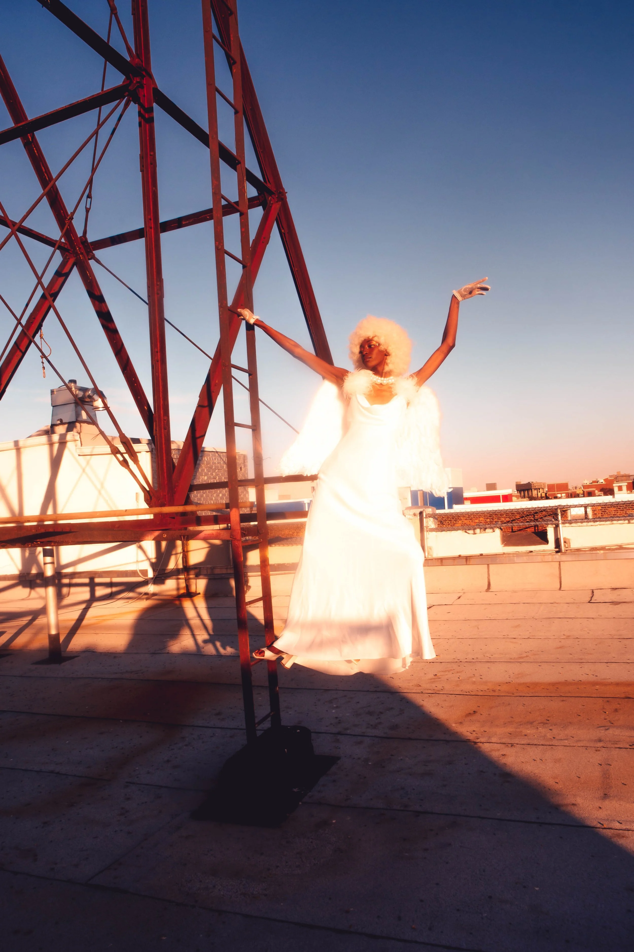 A person dressed in a white gown with a fluffy white cape and white gloves, posing with arms raised on a rooftop at sunset. There is a red metal tower structure and urban buildings in the background.