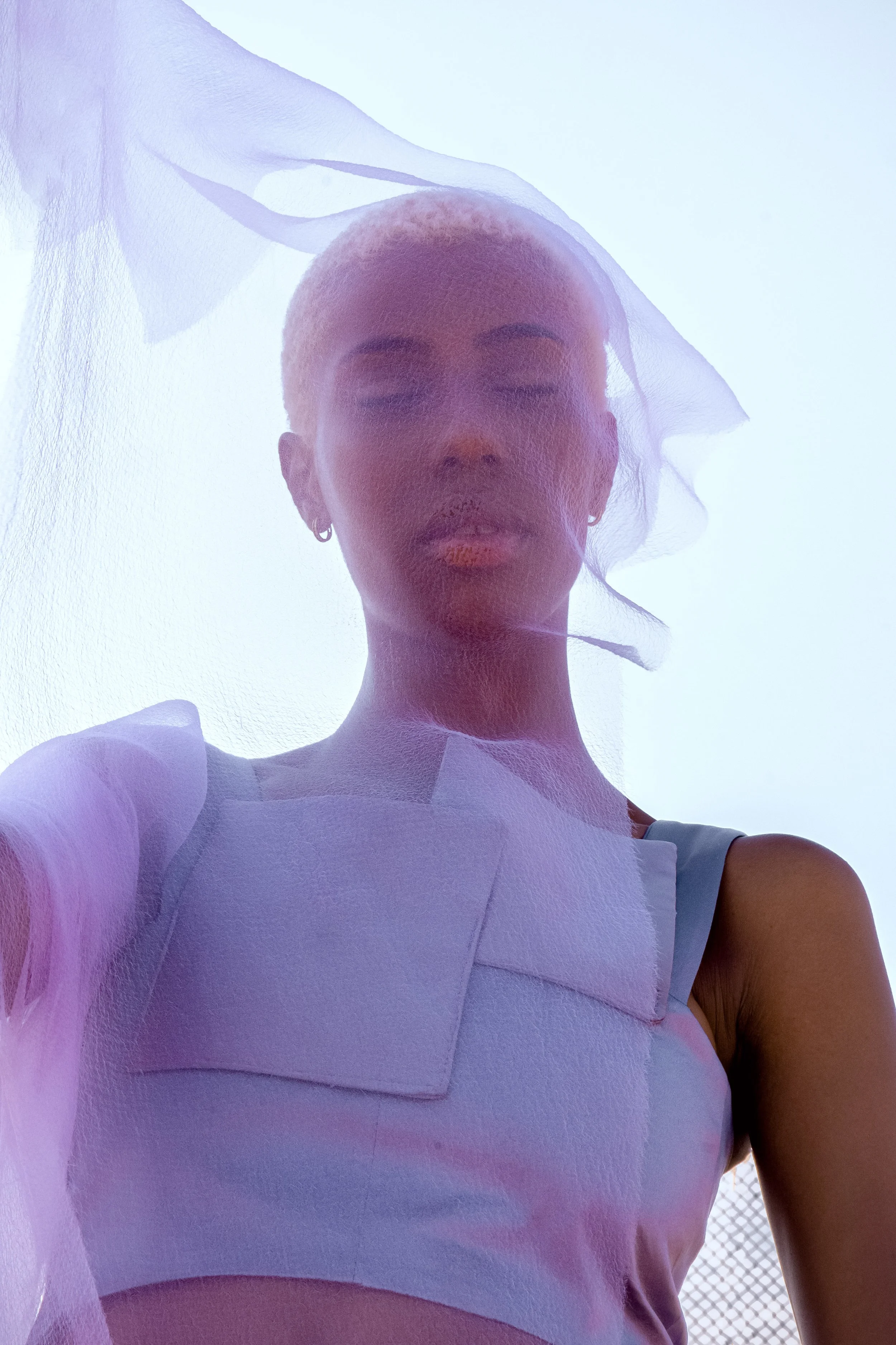 A woman with platinum blonde hair, earrings, and makeup wearing a white top, seen from below, with a sheer fabric draped over her head against a pale blue sky.