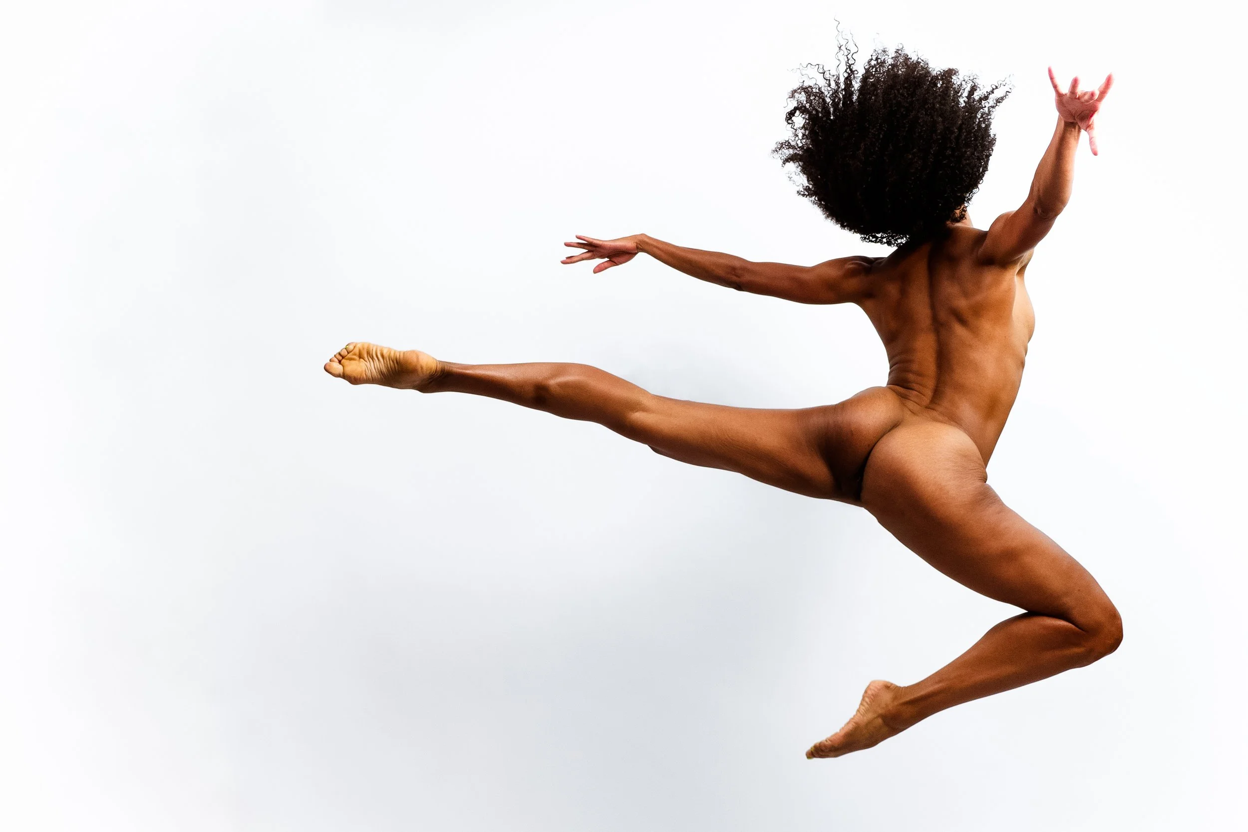 A person with curly hair and muscular build is performing a yoga pose against a plain white background, balancing on one leg with arms extended outward.