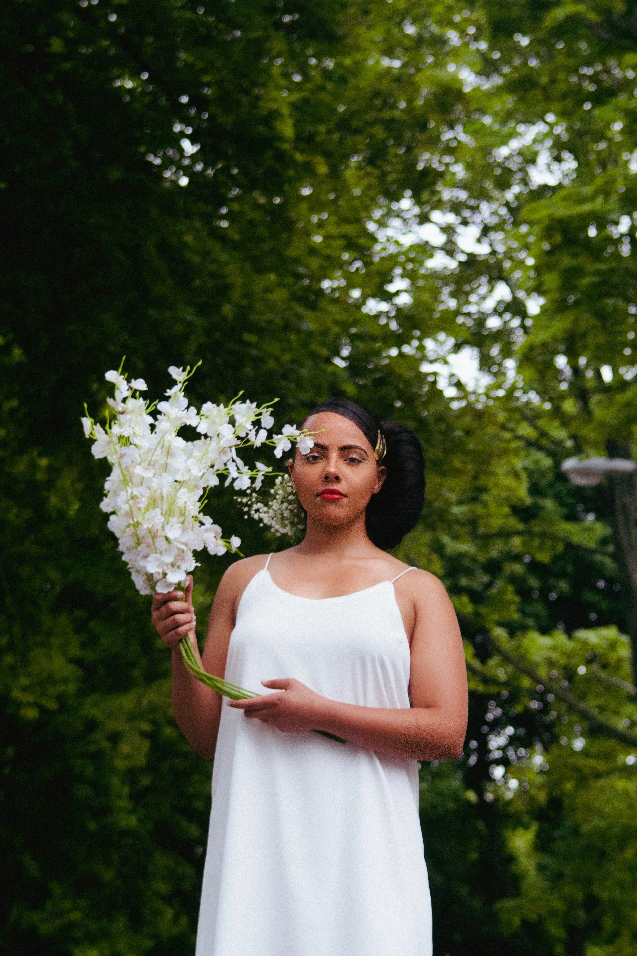 A woman with dark hair styled in an updo with hair accessories, wearing a white sleeveless dress, holding white flowers, standing outdoors with a green leafy background.