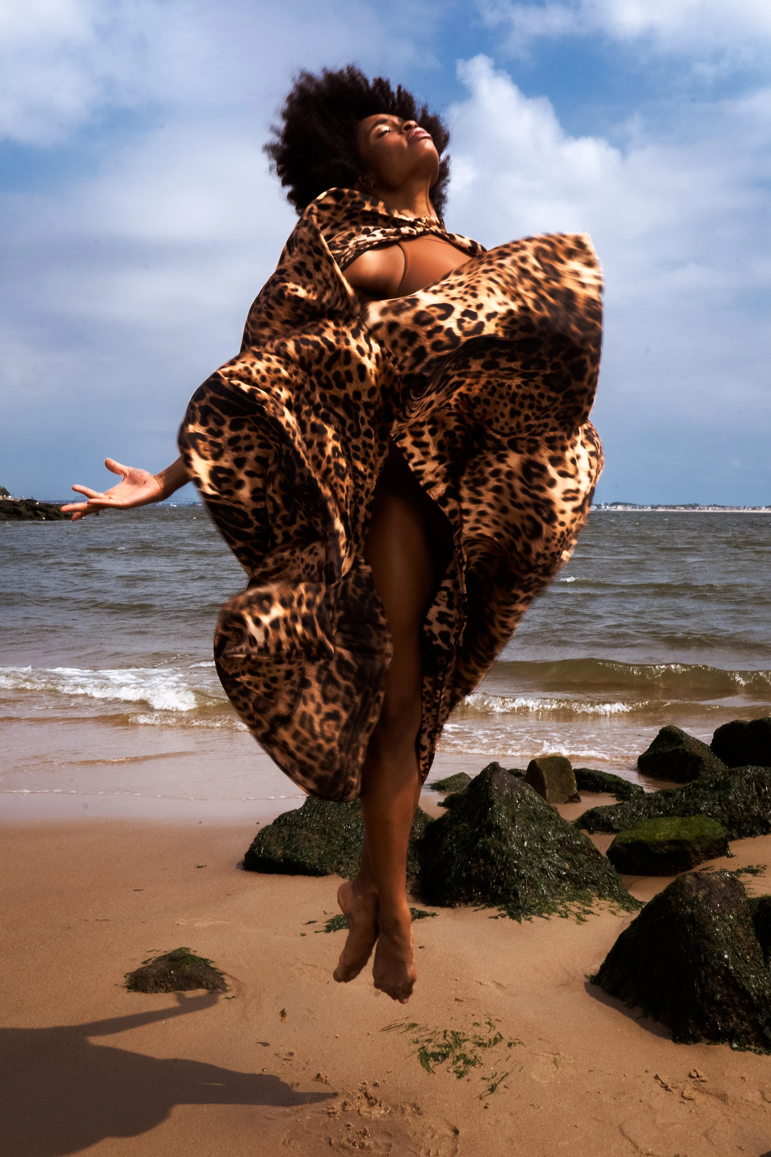 A woman standing on the beach with rocks, wearing a leopard-print robe, with her head tilted back, arms outstretched, and one leg raised, near the shoreline with the ocean and a cloudy sky in the background.
