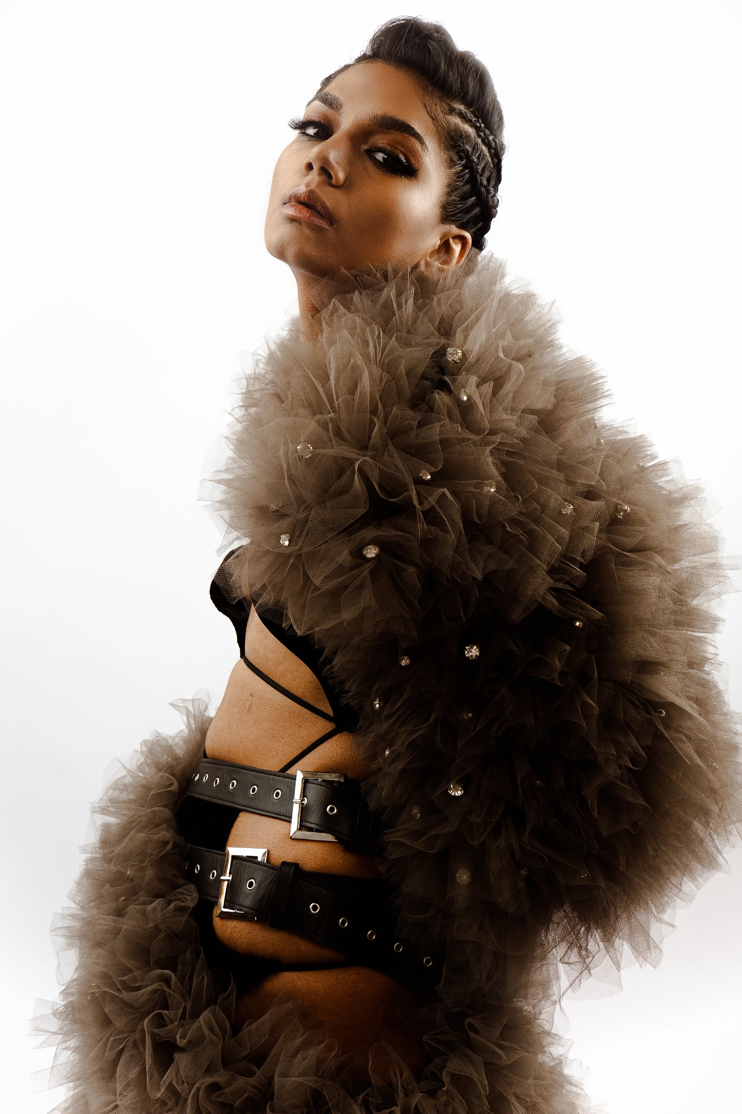 A woman with dark hair styled in braids, wearing smoky eye makeup and a nude lipstick, is wrapped in a large, ruffled, brown tulle fabric, adorned with small rhinestones. She has a confident expression and is wearing a black belt with metal buckles.