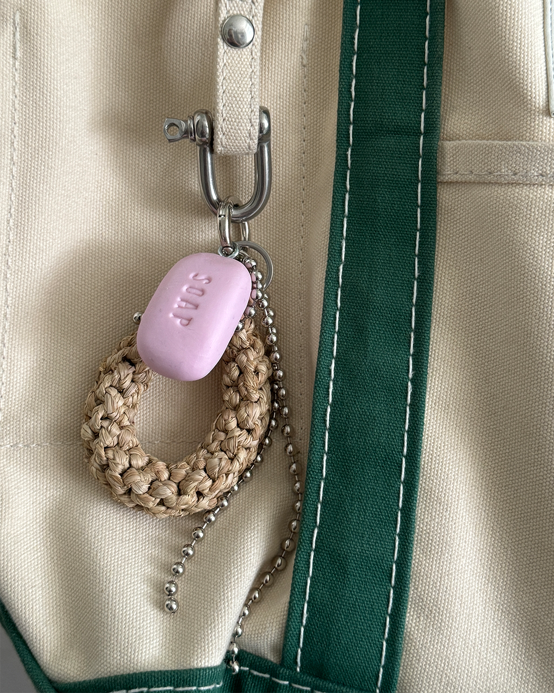 A beige and green bag with a metal hook holding a pink soap-shaped keychain, a woven beige loop, and a silver ball chain.