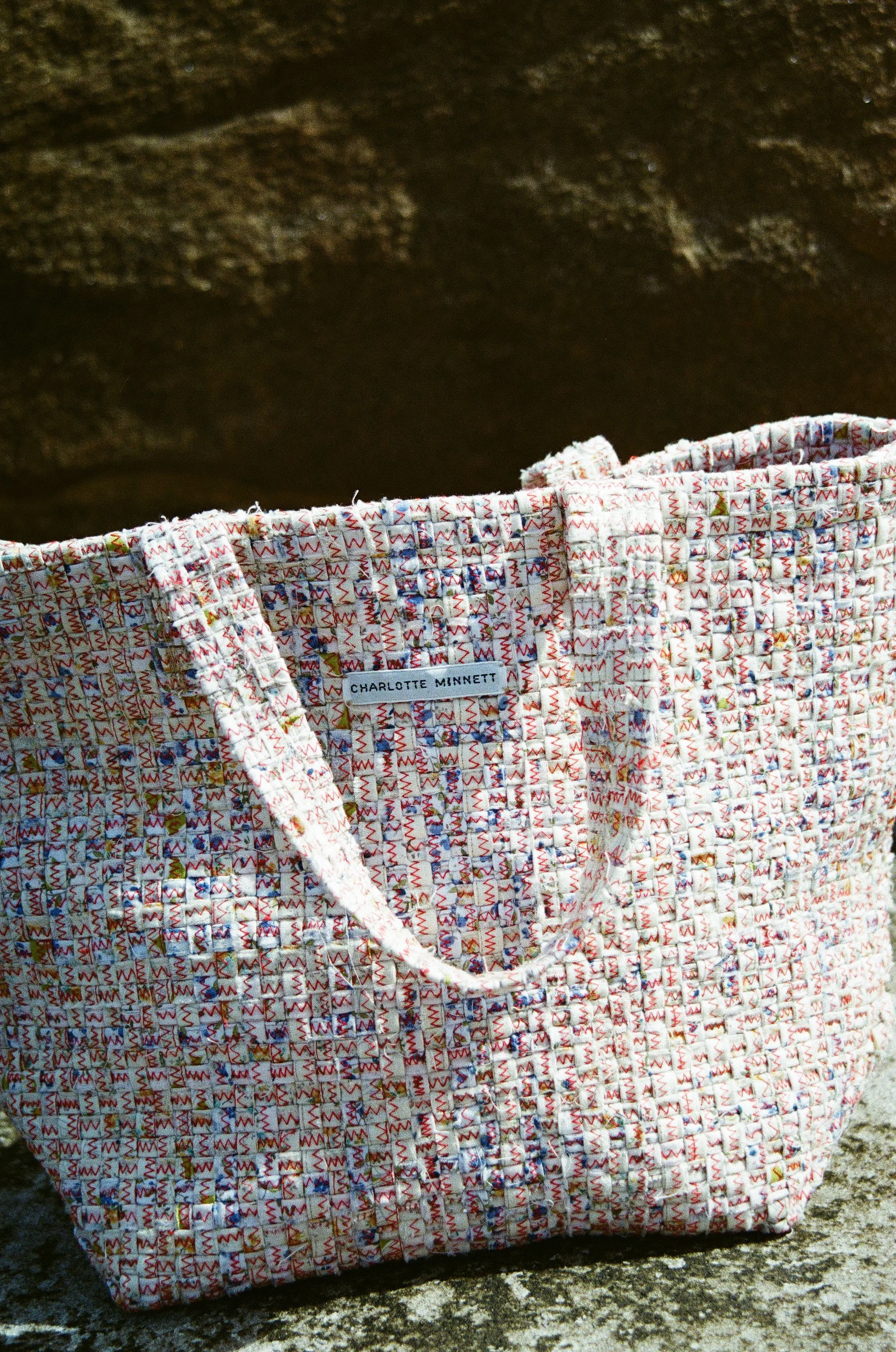 Colorful woven handbag with a Charlotte Minnett label placed on a textured stone surface.