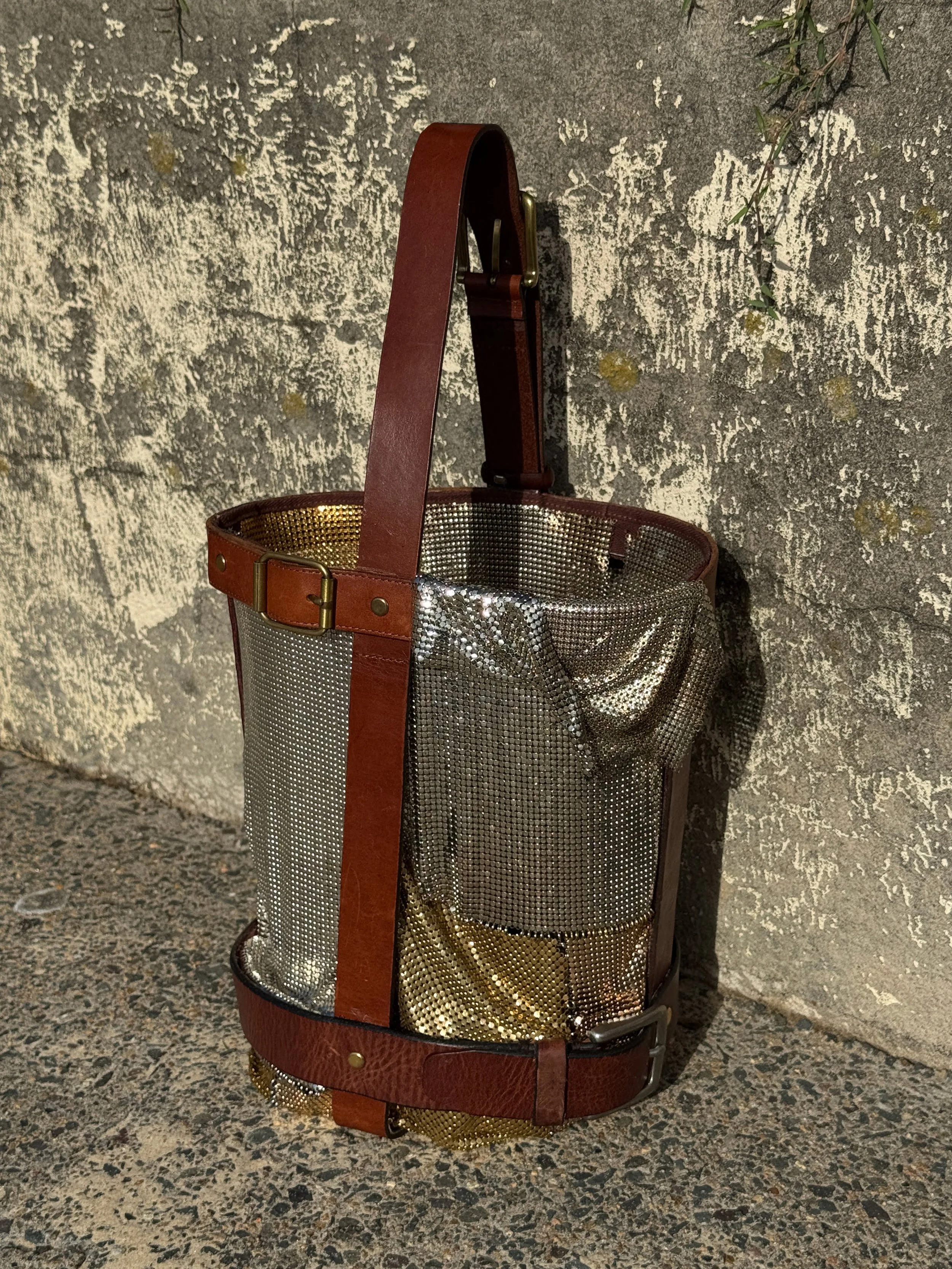 A metallic mesh basket with brown leather straps and handle, standing against a textured concrete wall.