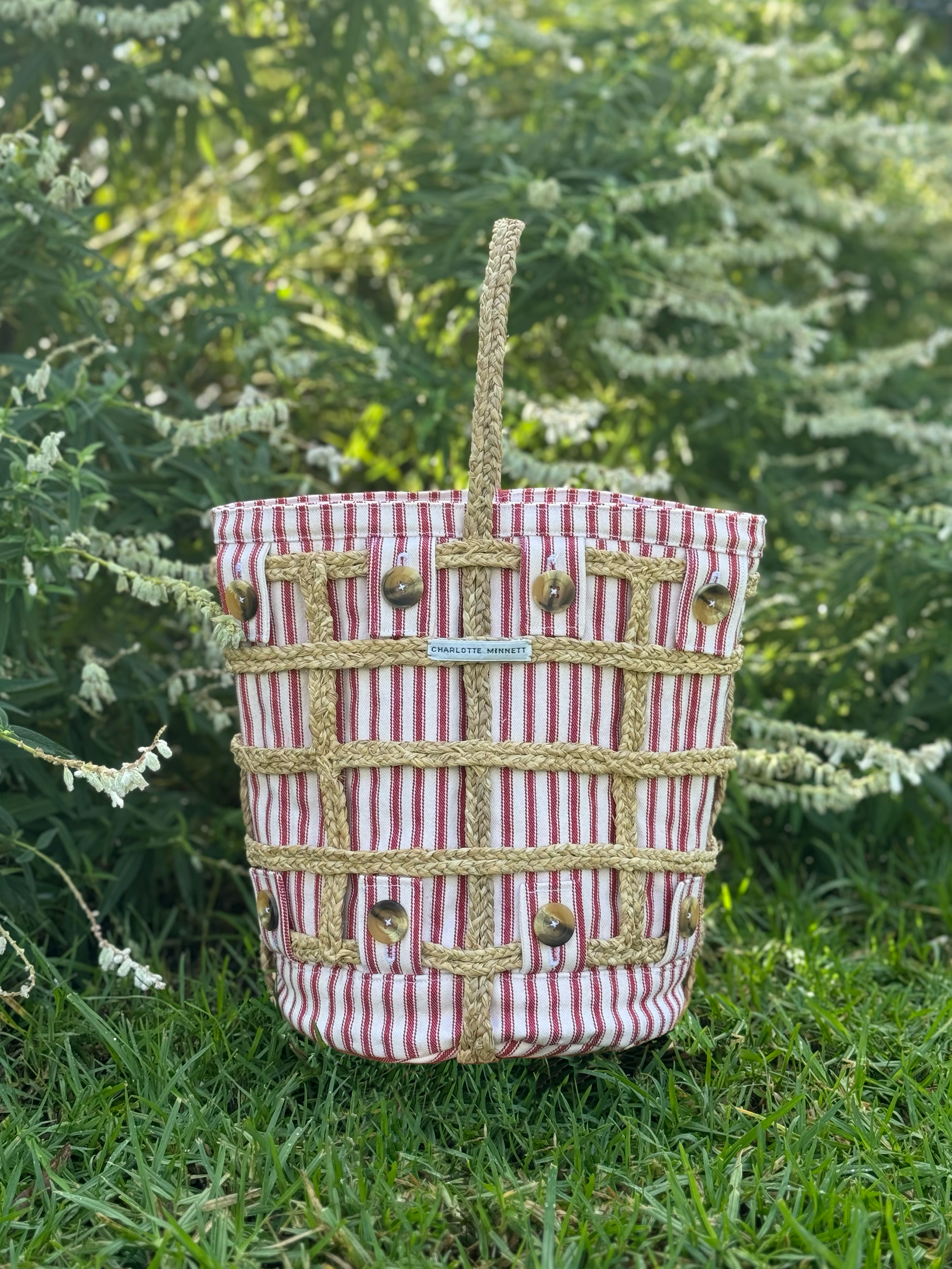 A woven straw handbag with a red and white striped fabric lining, decorated with round buttons and a woven handle, placed outdoors on grass with green foliage in the background.