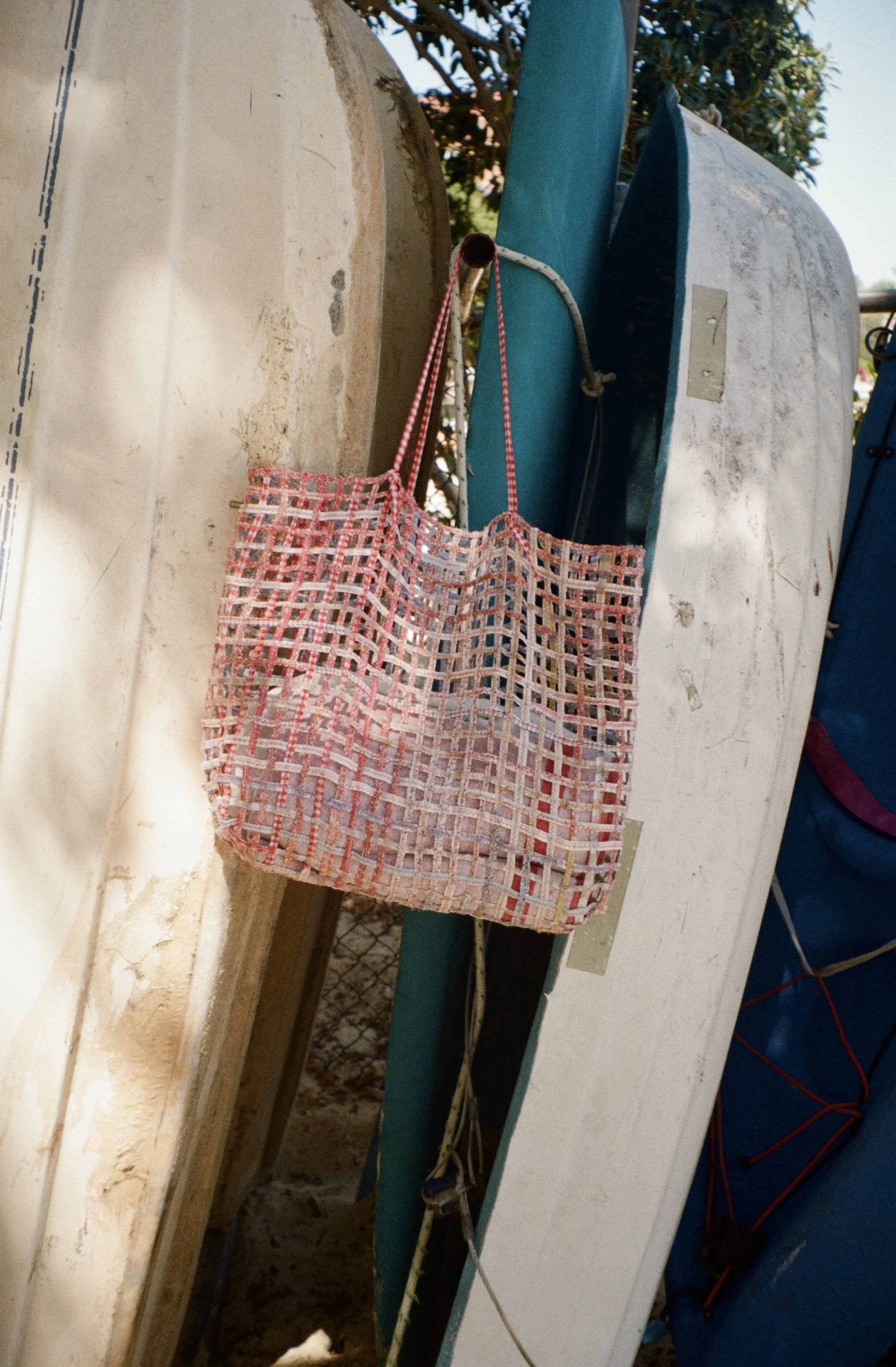 A pink woven tote bag hanging on a hook between boats with white and blue hulls, against a backdrop of a fence and trees.