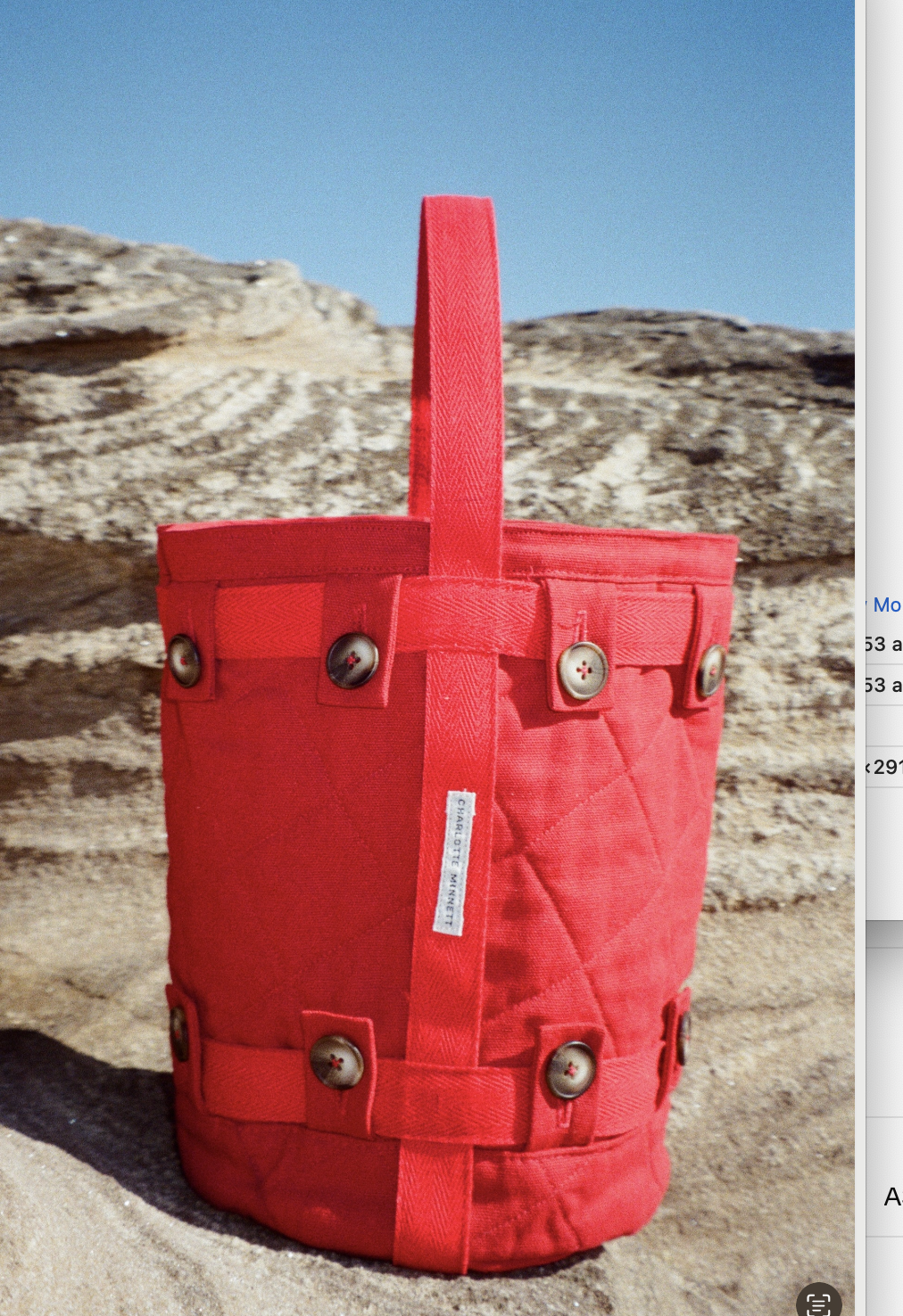 A red fabric tote bag with button details, placed on the sand in front of a rocky formation under a clear blue sky.