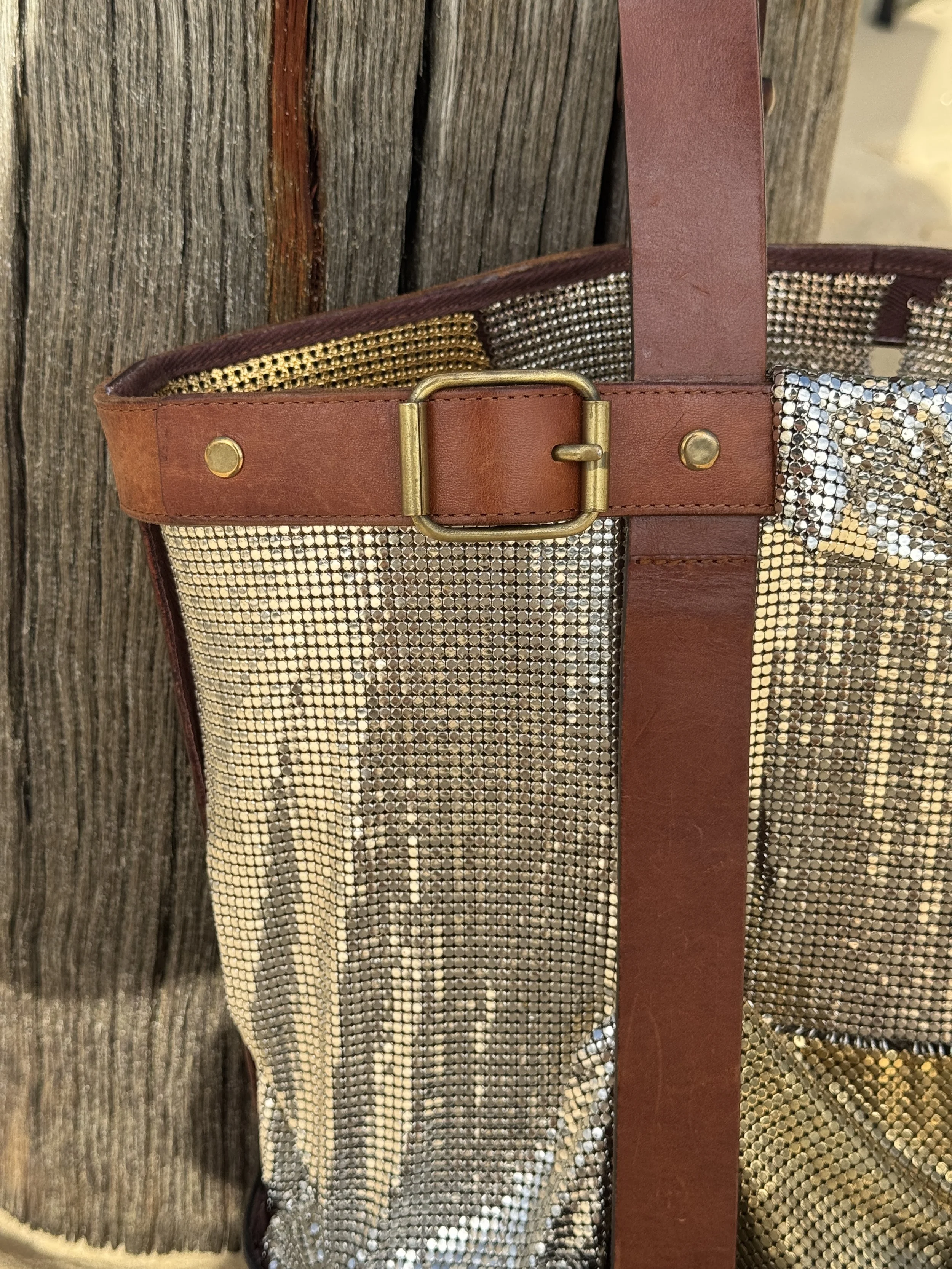 Close-up of a tote bag made with metallic mesh fabric, leather straps, and a leather belt with a brass buckle, leaning against a wooden surface.