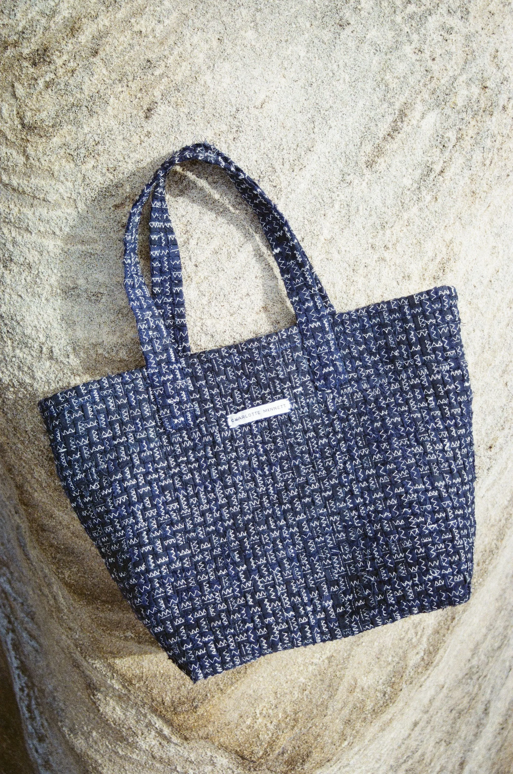 A navy blue tote bag with white zigzag and line patterns, lying on a sandy surface.