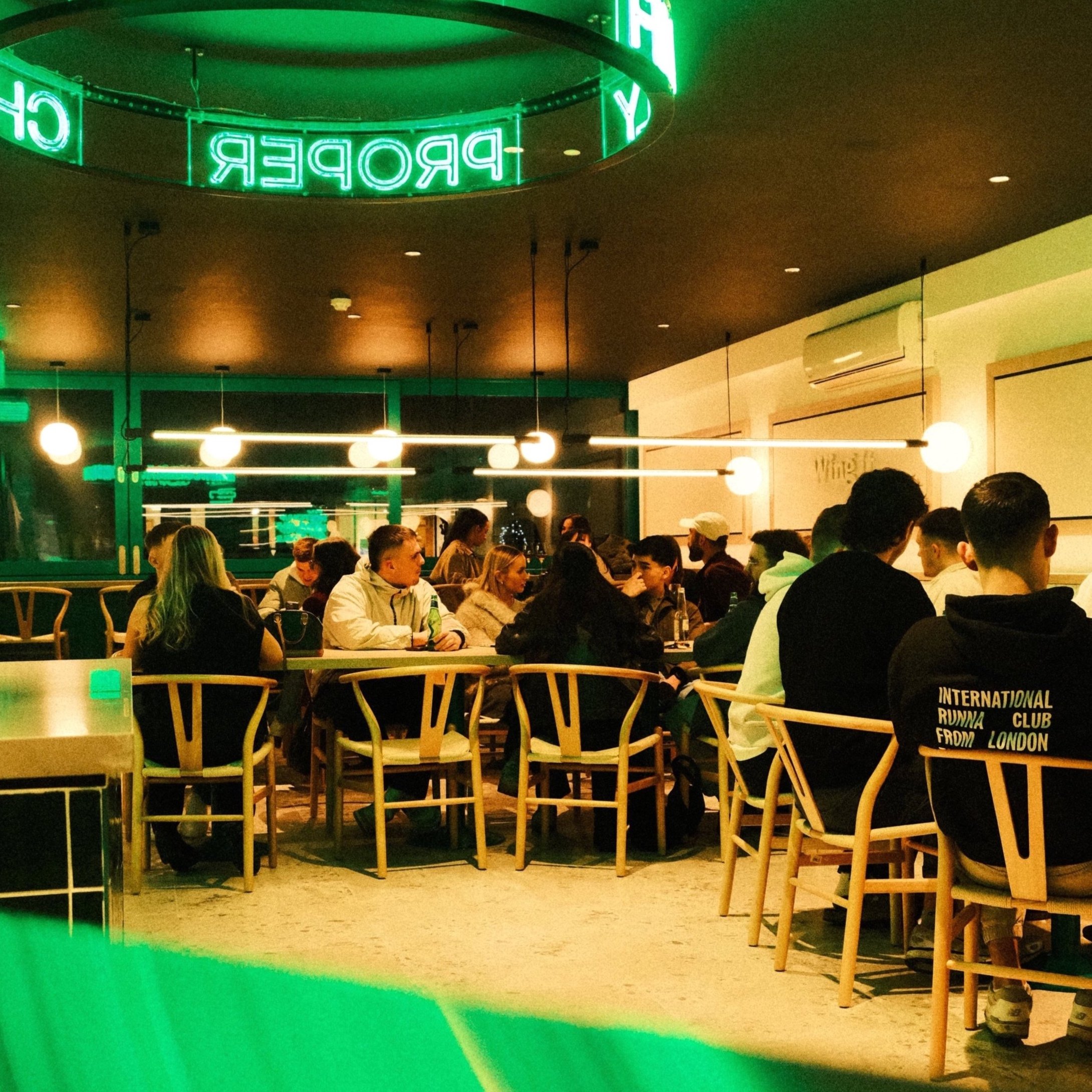 People dining at tables in a dimly lit restaurant with round hanging lights and green neon sign reading 'PROPER'