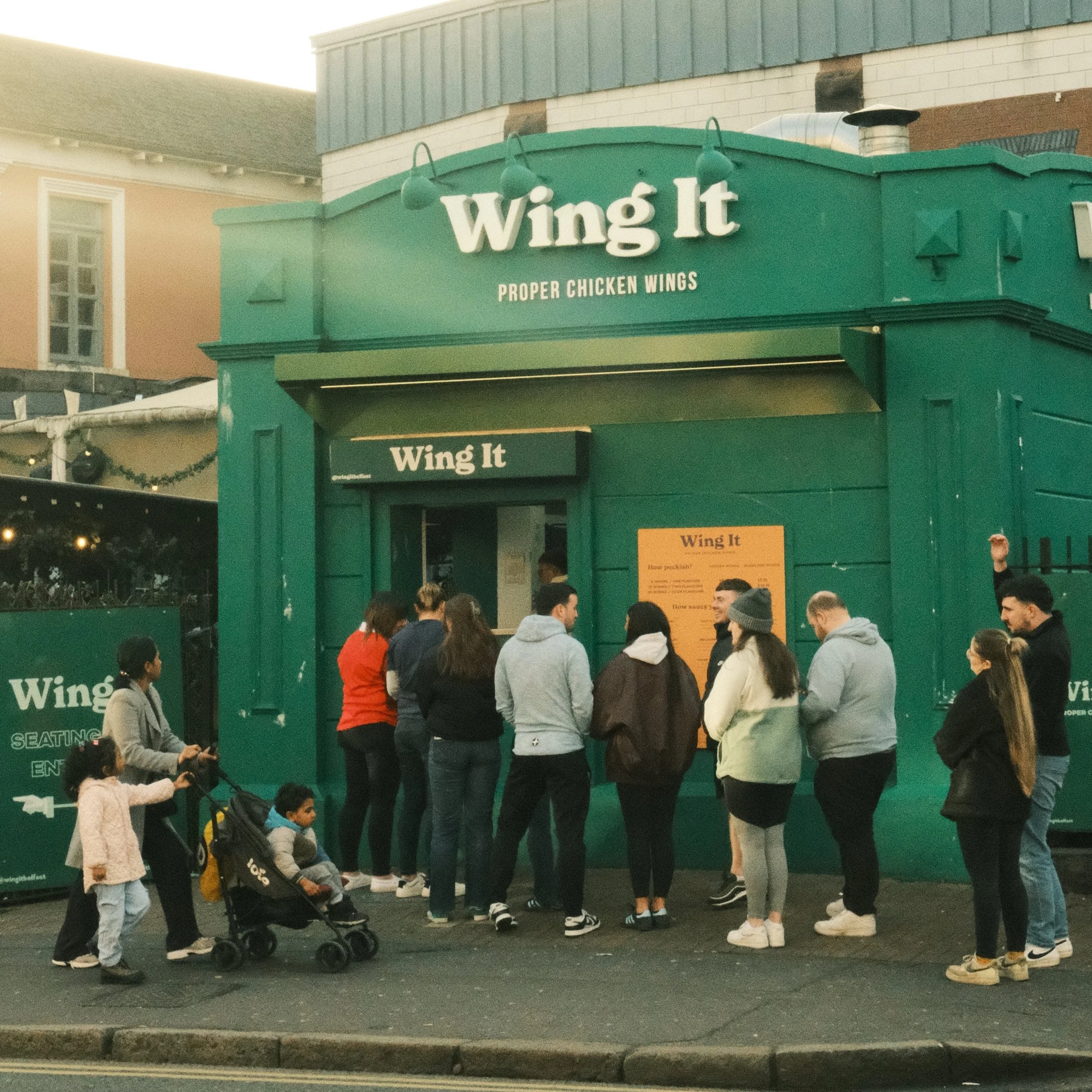 People standing in line outside a green food stand called 'Wing It' serving chicken wings.