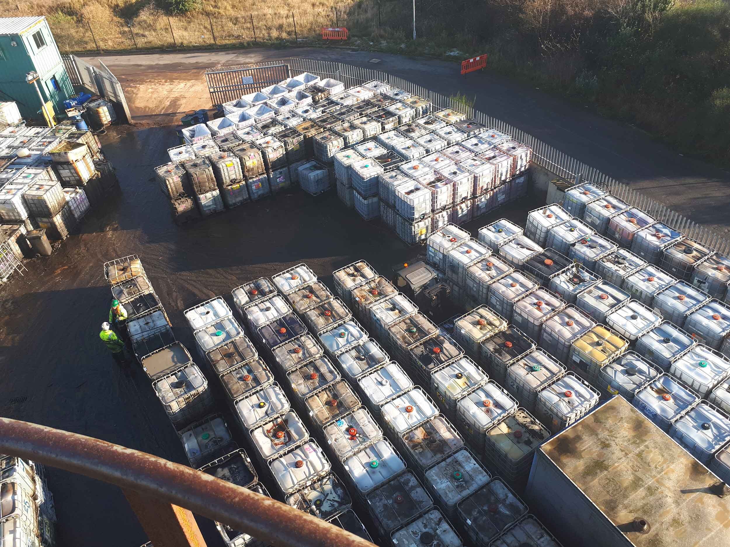 Rows of IBCs containing hazardous solid &amp; liquid wastes for treatment &amp; disposal with two people in high-vis jackets