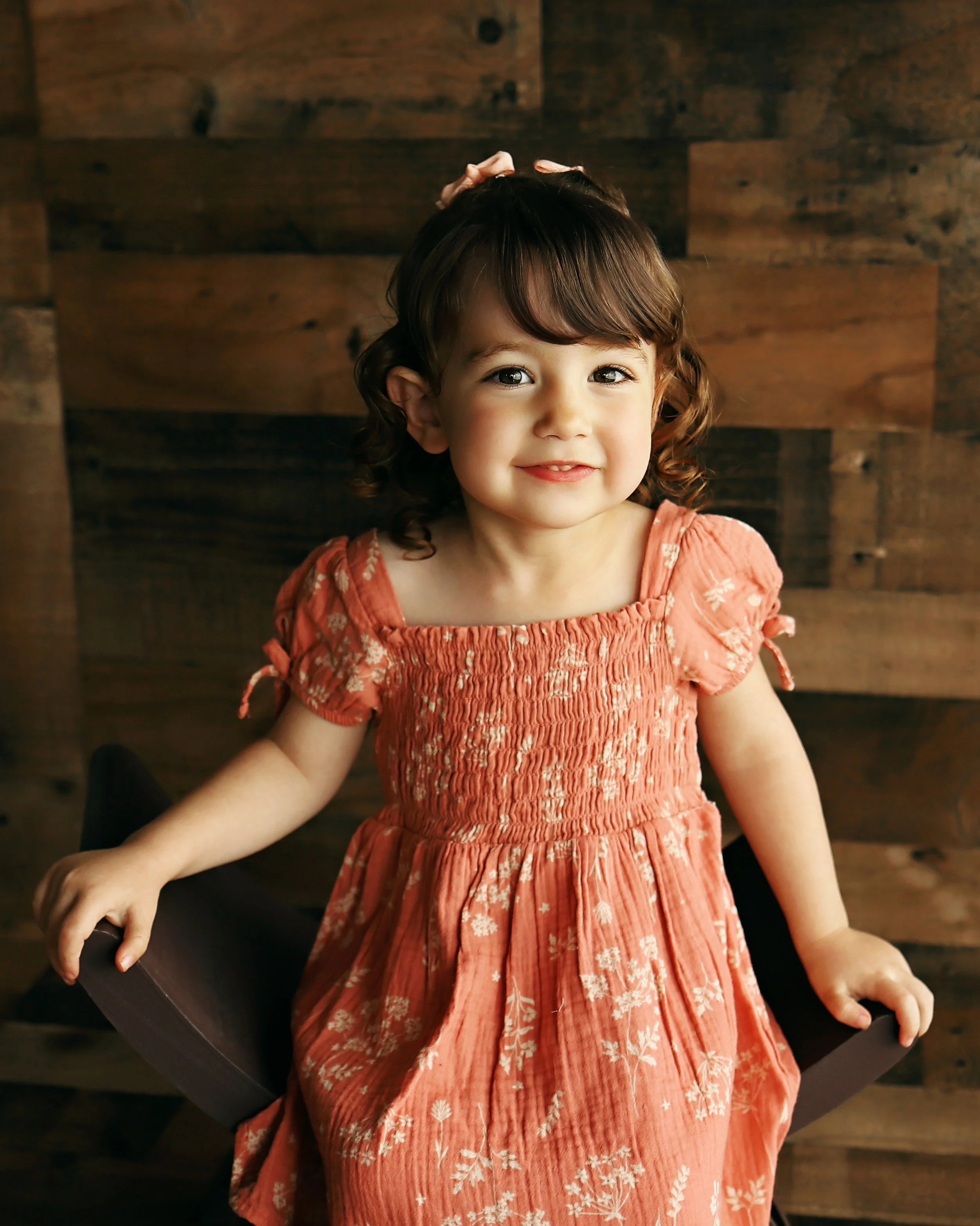 A young girl with curly brown hair and a smile, wearing an orange floral dress, sitting on a black chair in front of a wooden background.