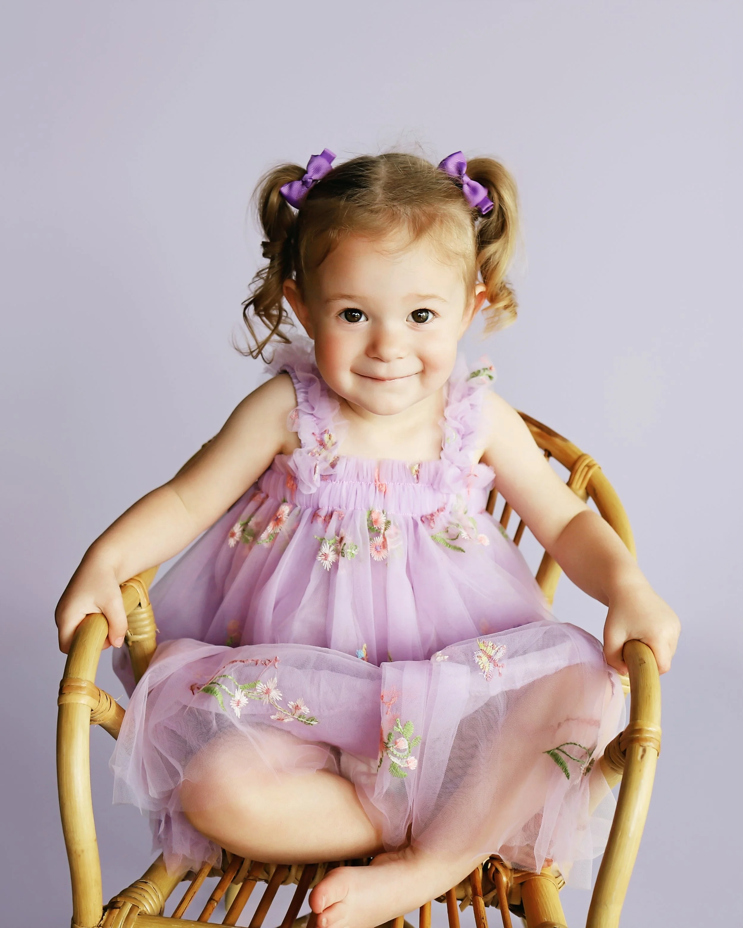 Smiling toddler girl in lavender floral dress sitting in a wicker chair during studio photo session in Christopher, Illinois