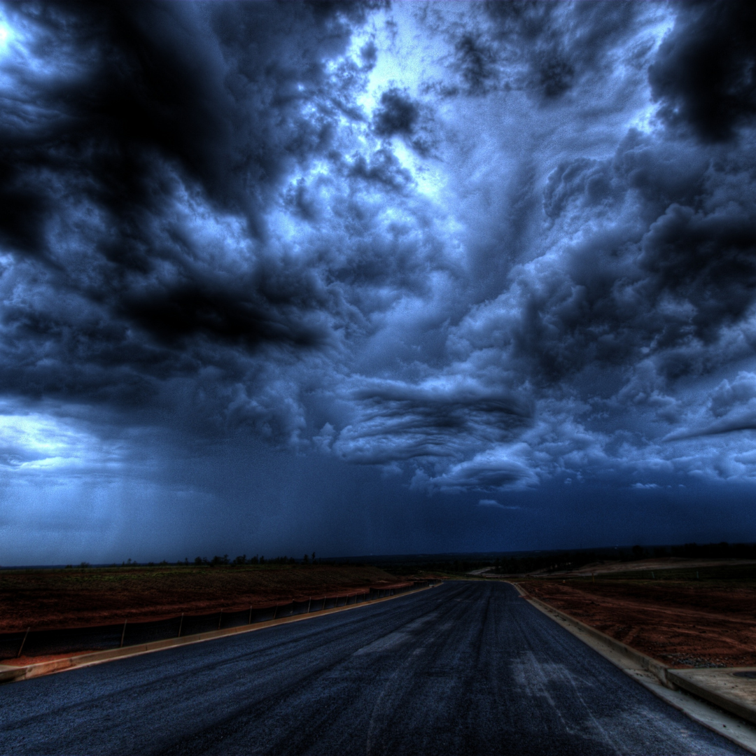 A dark, stormy sky with heavy clouds over an empty paved road in a rural area.
