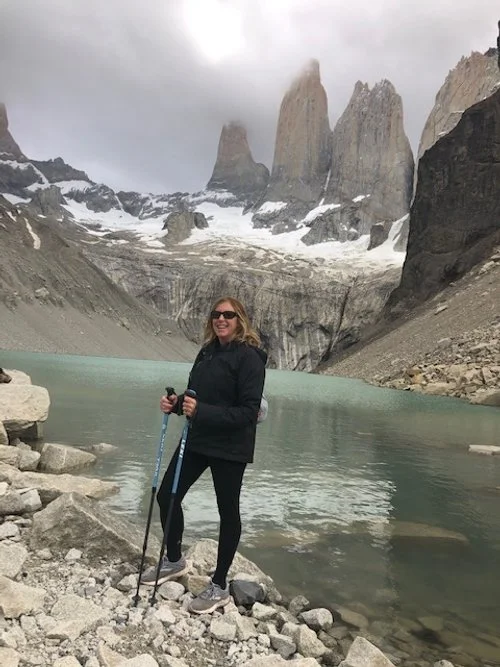 Woman with sunglasses smiling, holding hiking poles, standing on rocky shore near a lake with mountains and snow in the background.