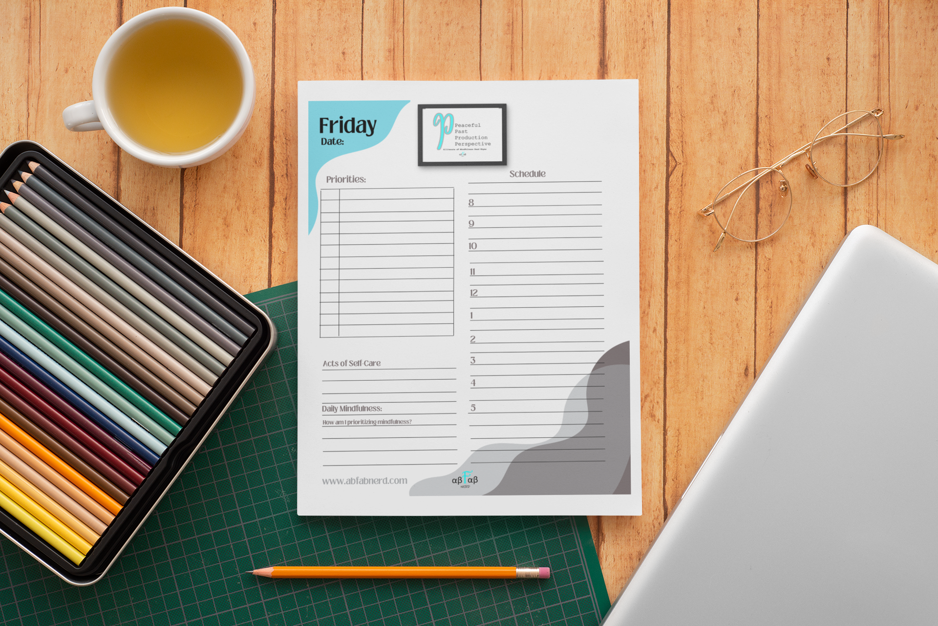 A wooden desk with a cup of tea, a colored pencil set in a black case, a green cutting mat, a downloadable planner sheet, a pair of gold glasses, and a silver laptop.