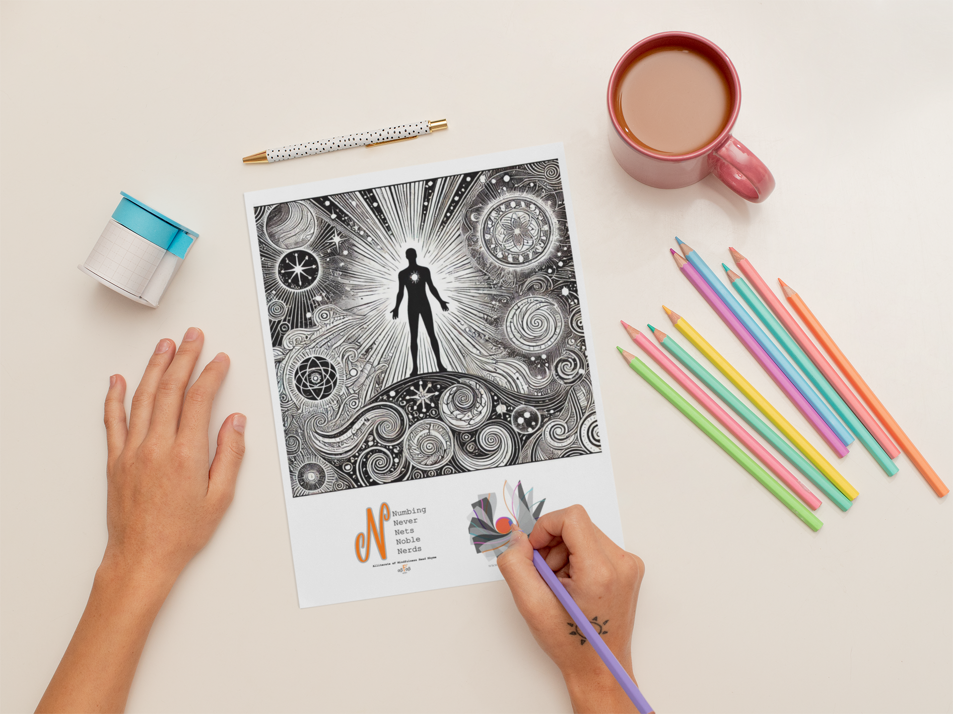 A person coloring a black-and-white intricate illustration of a human figure surrounded by celestial symbols. On the desk are colored pencils, a pink mug of tea, a decorative tape dispenser, a patterned pen, and a printed alphabet reference. The person's left hand rests on the desk, and right hand uses a purple colored pencil to color the illustration.