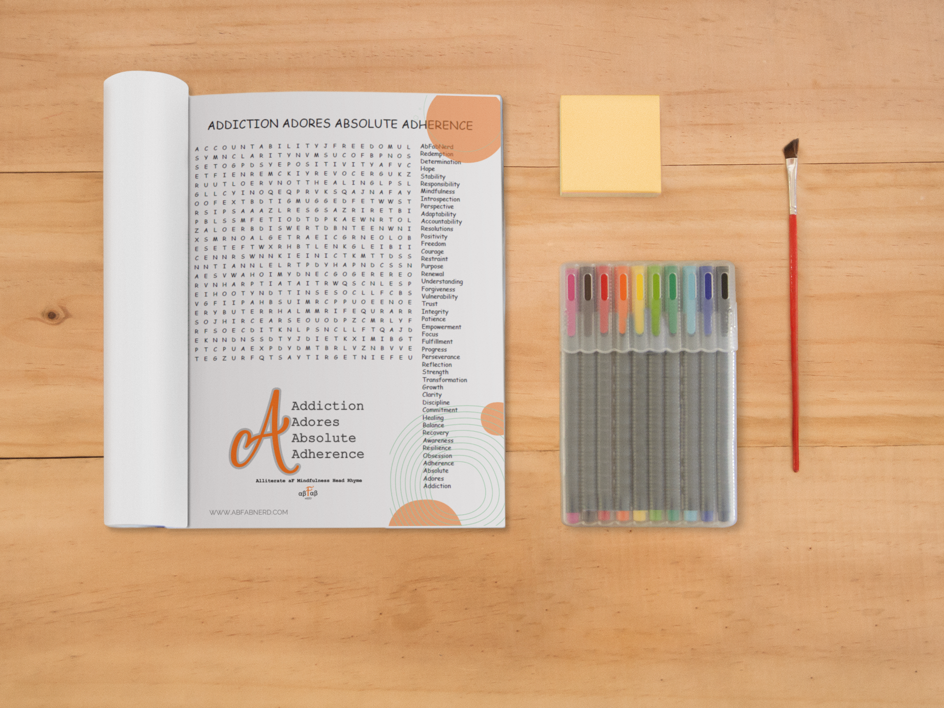 A flat lay of a word search puzzle titled "Addiction Adores Absolute Adherence" on white paper, a set of colored markers in a clear case, a yellow sticky note, and a paintbrush on a wooden surface.