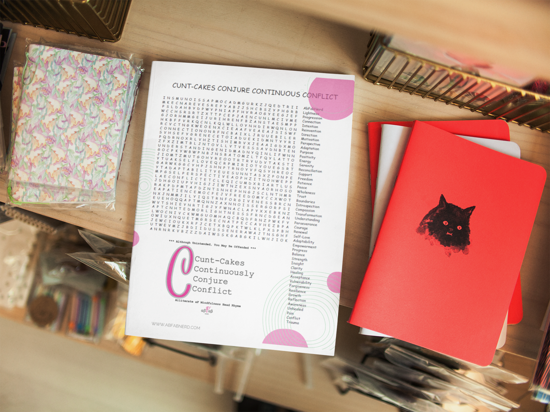A desk with a word search puzzle titled 'Cunt-Cakes Conjure Continuous Conflict,' a small pink sticker, some blank notebooks with a red cover and a black cat illustration, and a clear plastic bag with colorful patterned items inside.