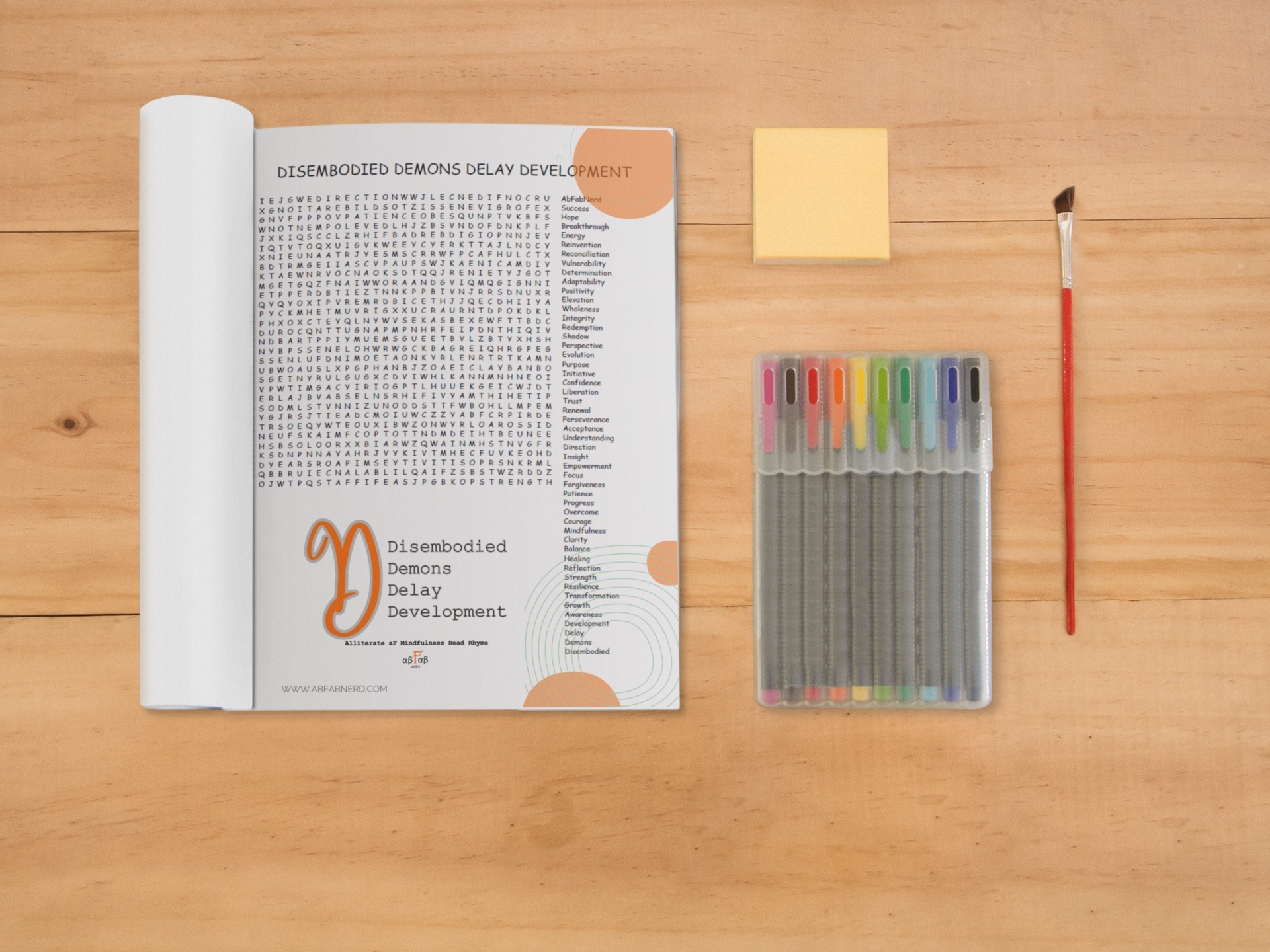 A table with a book titled 'Disembodied Demons Delay Development' open to a page with a word search puzzle, a small stack of yellow sticky notes, a set of colorful gel pens in a case, and a red-handled paintbrush.