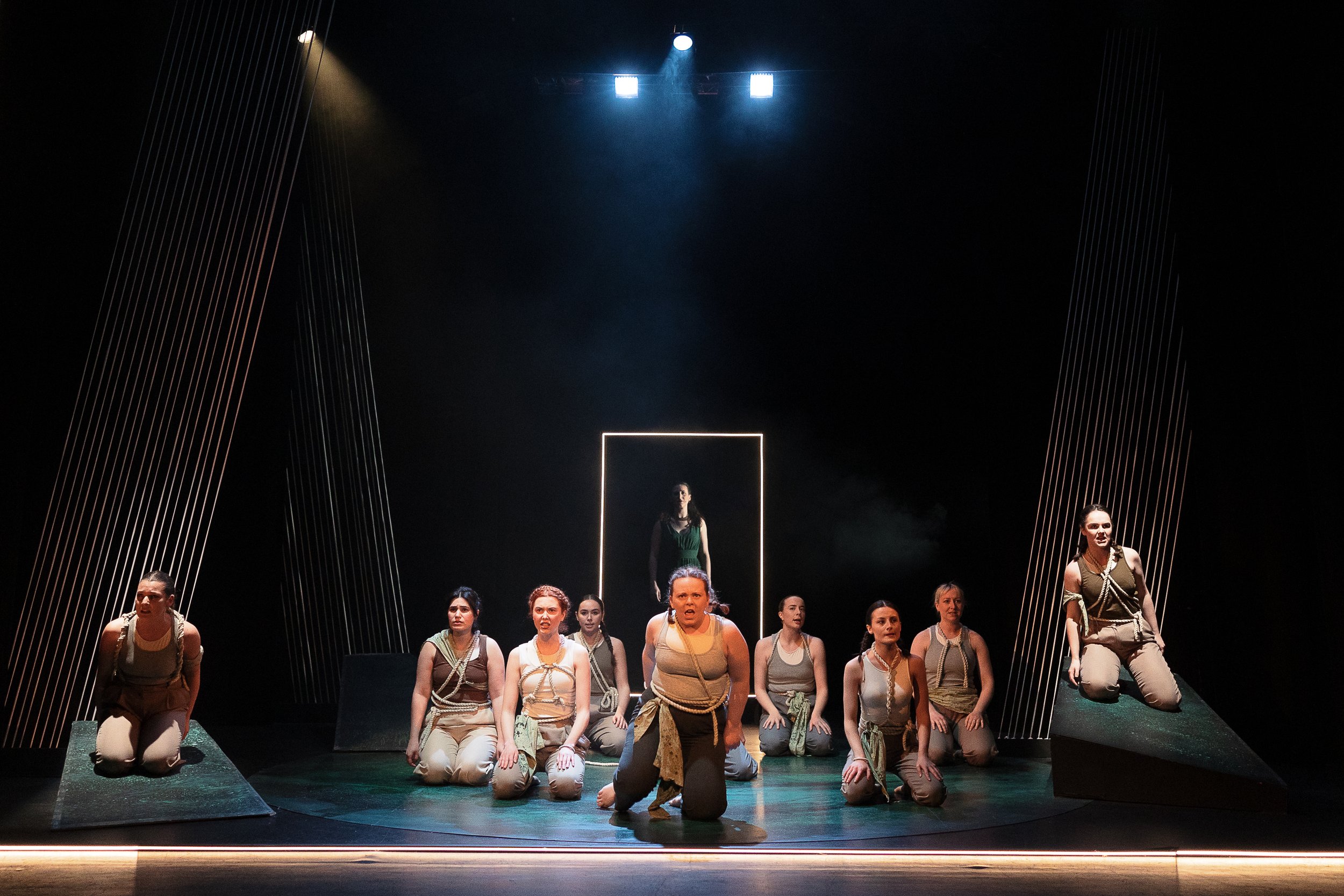 Theatrical performance with a group of actors on stage, dressed in coordinated costumes. The scene is minimally lit, with dramatic lighting focusing on the actors. The background features a lit rectangular frame and diagonal lines, suggesting a moder