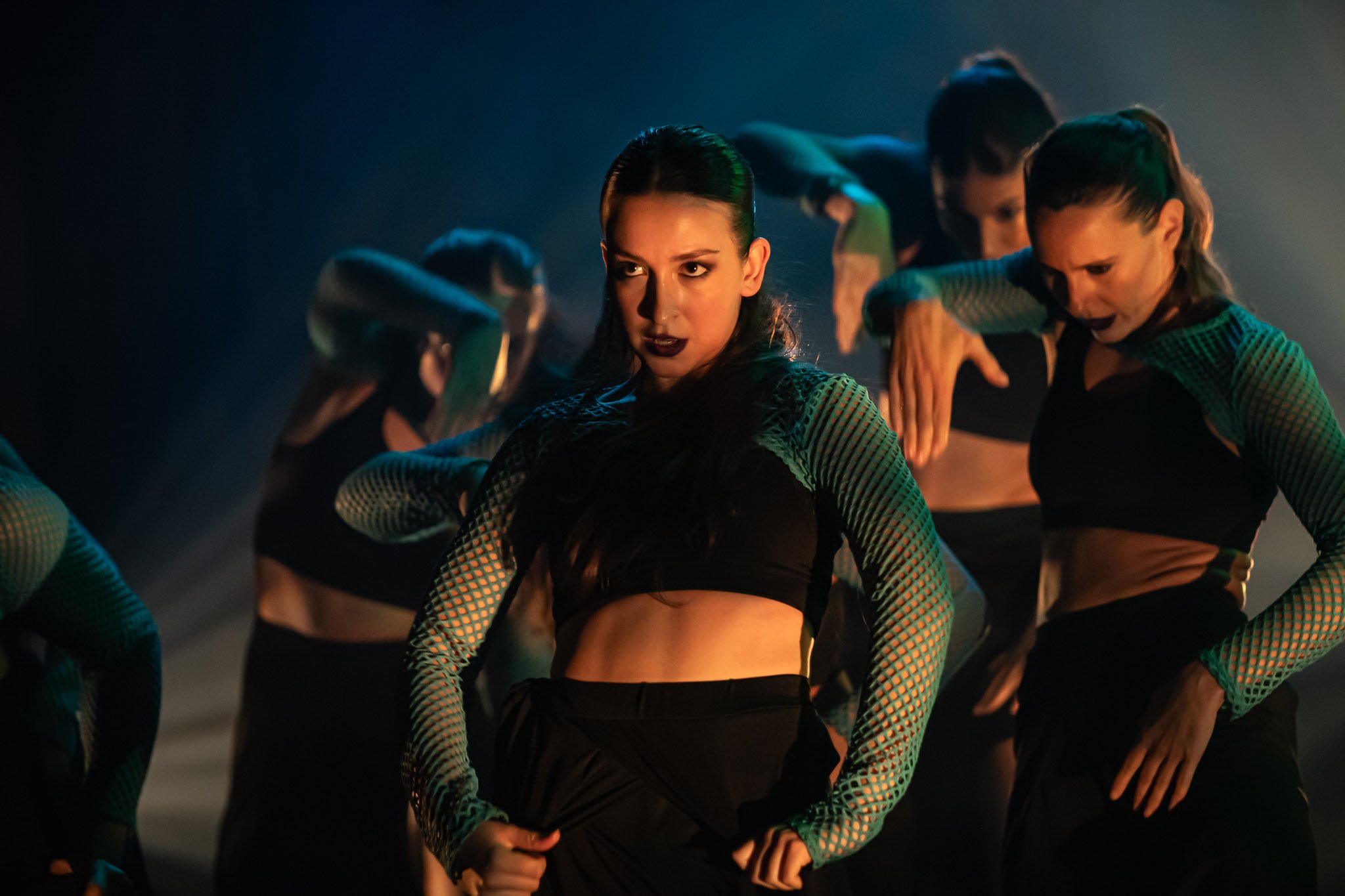 Group of dancers performing on stage with dramatic lighting, wearing dark outfits with mesh sleeves.