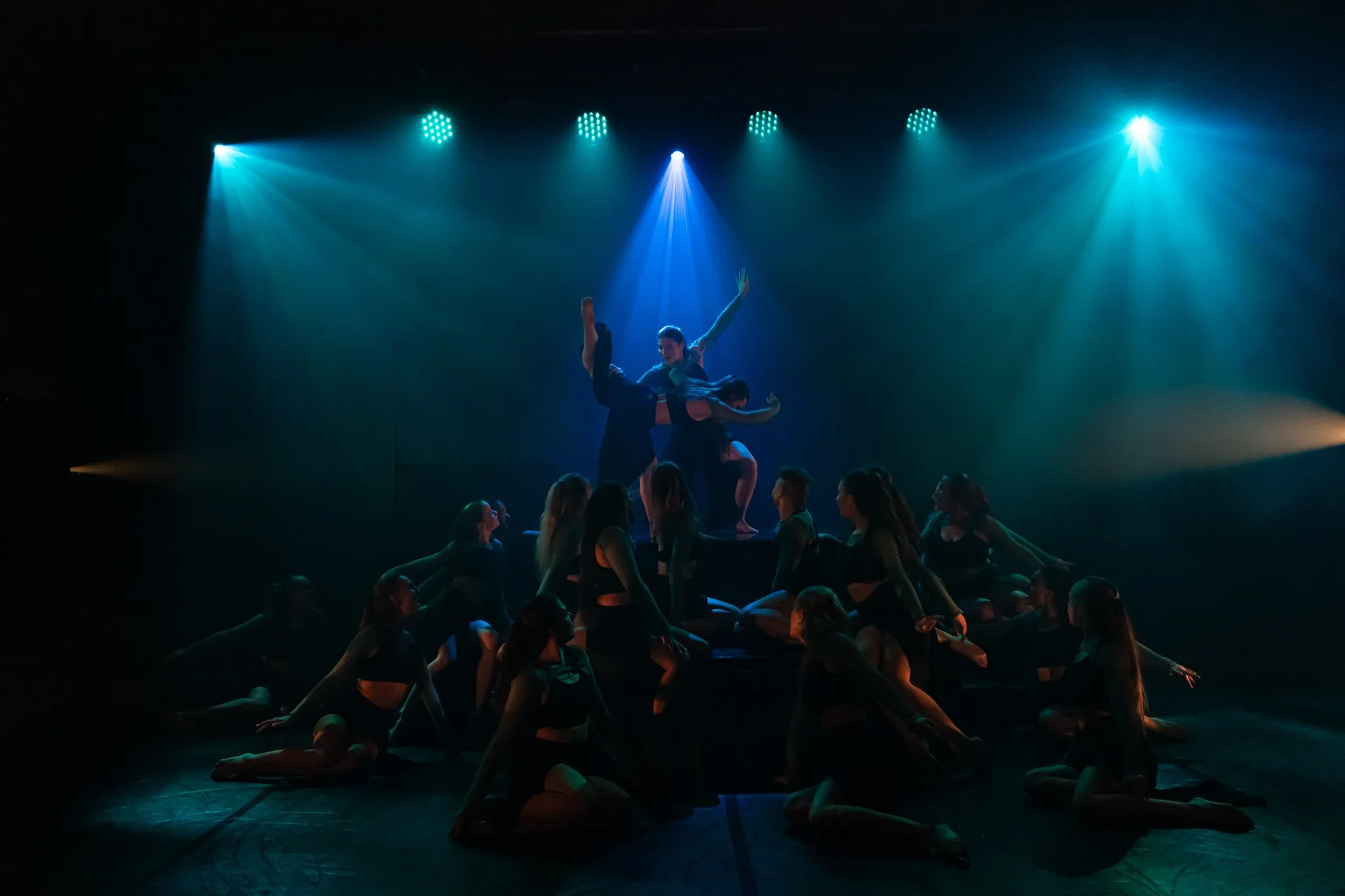 Dance performers on stage in dim blue lighting, with one dancer elevated in the center, surrounded by others sitting and posing.