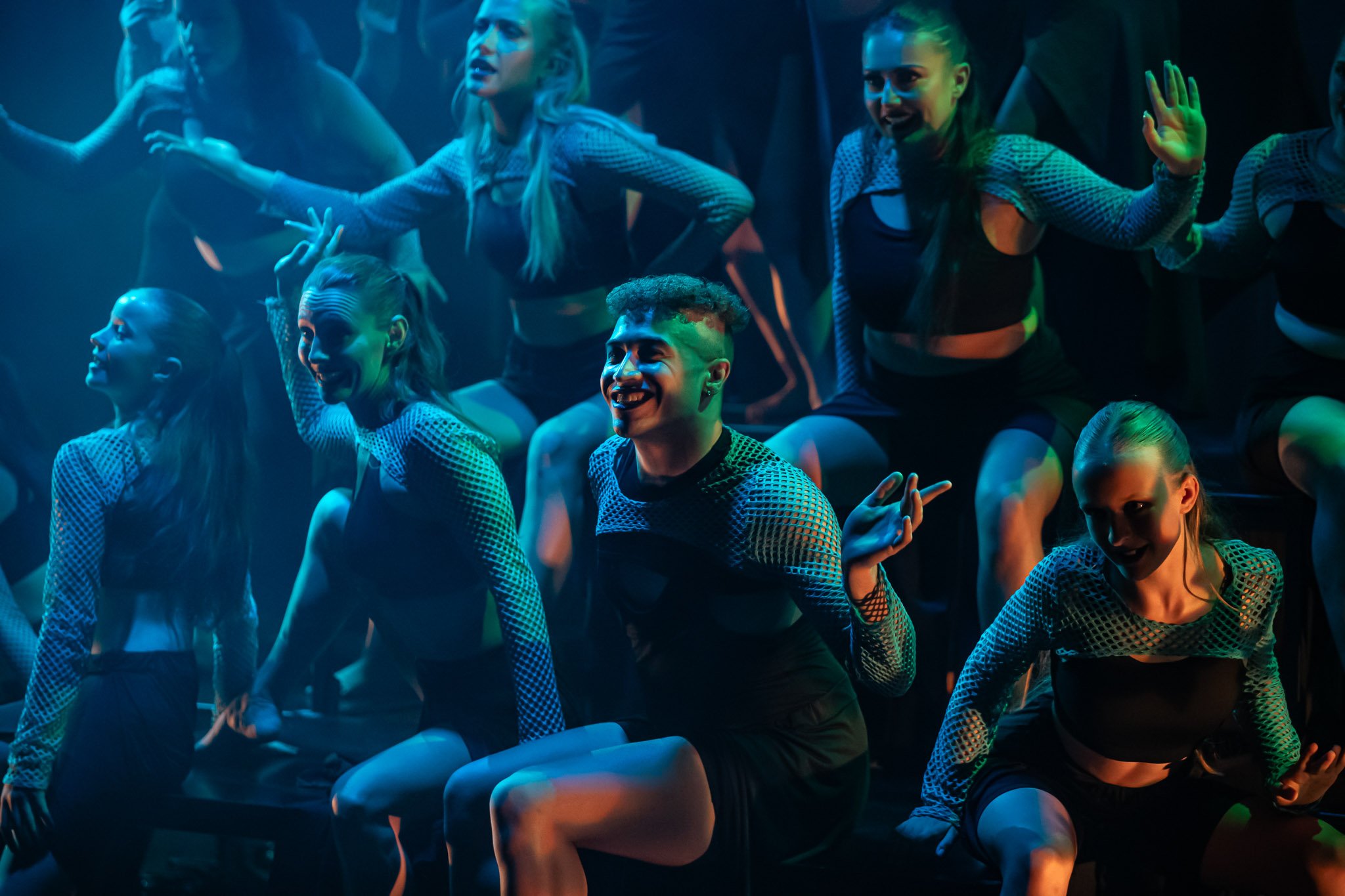 Group of dancers performing on stage with expressive poses, wearing mesh tops and shorts, in colorful theatrical lighting.